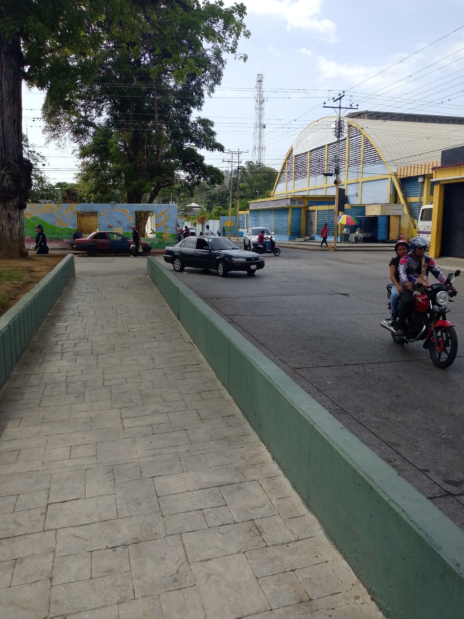 The image captures a vibrant daytime street scene in Tinaquillo, Venezuela, featuring a mix of urban infrastructure, vehicular traffic, and pedestrians.
In the foreground, a textured gray-paved walkway, bordered by green concrete curbs, leads into the frame. To the left, large green trees provide ample shade, and behind them, a colorful mural depicting birds in flight over a landscape adorns a wall. Several people are visible near this mural, including one in a dark uniform kneeling, and others standing or walking. Two cars are on the road in this area: a two-toned sedan (red and dark blue/black) and a black sedan.
The asphalt road occupies the midground and right side of the image. A red motorcycle, carrying two riders (a helmeted driver in protective gear and a female passenger), is prominently featured on the right, heading towards the viewer. Further down the street, another motorcycle and a white van are visible.
A distinctive large building with a yellow-arched roof and blue and yellow walls dominates the right background. Text visible on this building identifies it as "GIMNASIO CUBIERTO PEDRO CONTRERAS" (Pedro Contreras Covered Gymnasium). Power lines crisscross the sky above the building. In the distant left background, a tall telecommunications tower stands against a bright, partly cloudy sky.
The scene depicts everyday activity with people commuting and going about their routines under what appears to be clear, fair weather conditions.