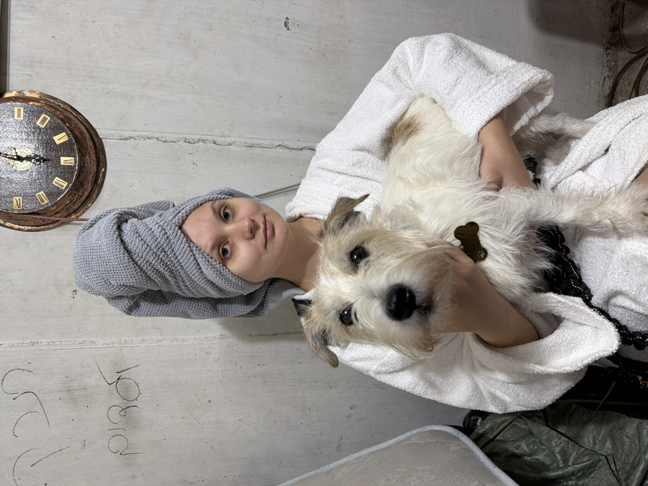 A young adult female is depicted holding a small, white terrier-type dog with brown markings on its ears and back. The woman, with fair skin and light-colored eyes, has her hair wrapped in a grey, textured towel turban and wears a white terry cloth bathrobe, partially revealing a black lace garment underneath. Both the woman and the dog gaze directly forward, towards the camera. The dog wears a dark, bone-shaped tag on its collar.
The background consists of a rough, light grey concrete wall. Prominently displayed on the wall to the left of the woman's head are two lines of handwritten Hebrew graffiti: the top line reads "תכף" (Tēkef, meaning "soon" or "shortly"), and the bottom line reads "לגגות" (Lagagōt, meaning "to the roofs"). Above and to the right of the woman's head, a circular, hammered copper-toned wall clock with Roman numerals indicates a time of approximately 12:35. To the far left, a white mattress or cushion leans against the wall, partially covered by a dark tarp or fabric.
The setting, characterized by the unfinished concrete walls and the specific Hebrew graffiti "תכף לגגות," strongly suggests a repurposed underground space or bomb shelter environment in Tel-Aviv, Israel, a location often associated with specific cultural venues like Rothschild 10, known for its unique interior design elements and connection to rooftop spaces.
