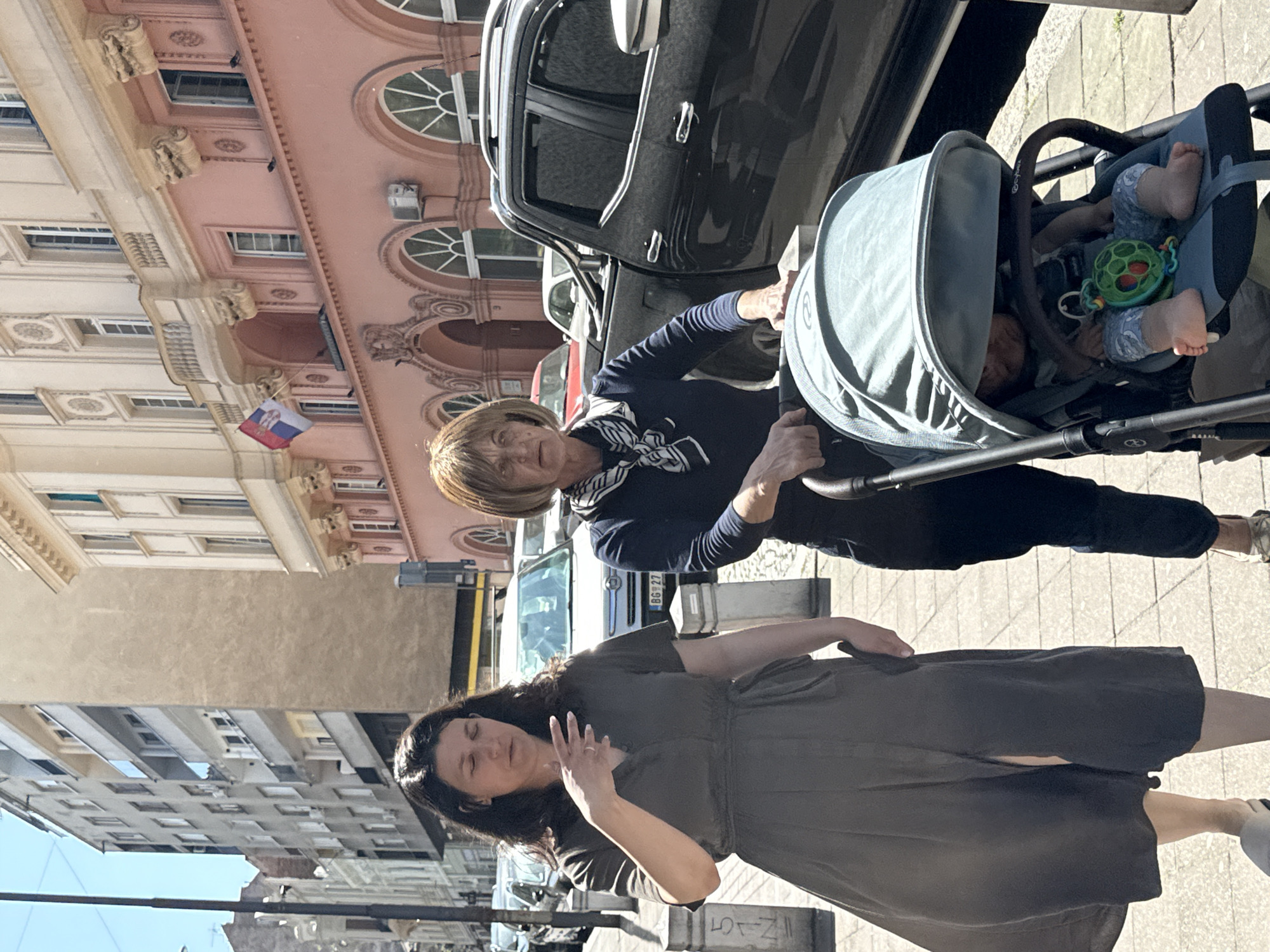 The image depicts two women on a city street in Belgrade, Serbia. One woman, who appears older with short blonde hair, is pushing a stroller containing a baby. The baby is lying down, with its legs visible and a toy nearby. The older woman is wearing a dark jacket and a patterned scarf. A black SUV is parked next to them. The second woman, with long dark hair, is wearing a dark dress and has her hand raised, as if gesturing or posing. Buildings line the street, including one with a Serbian flag. The architectural style suggests a historic area of the city.