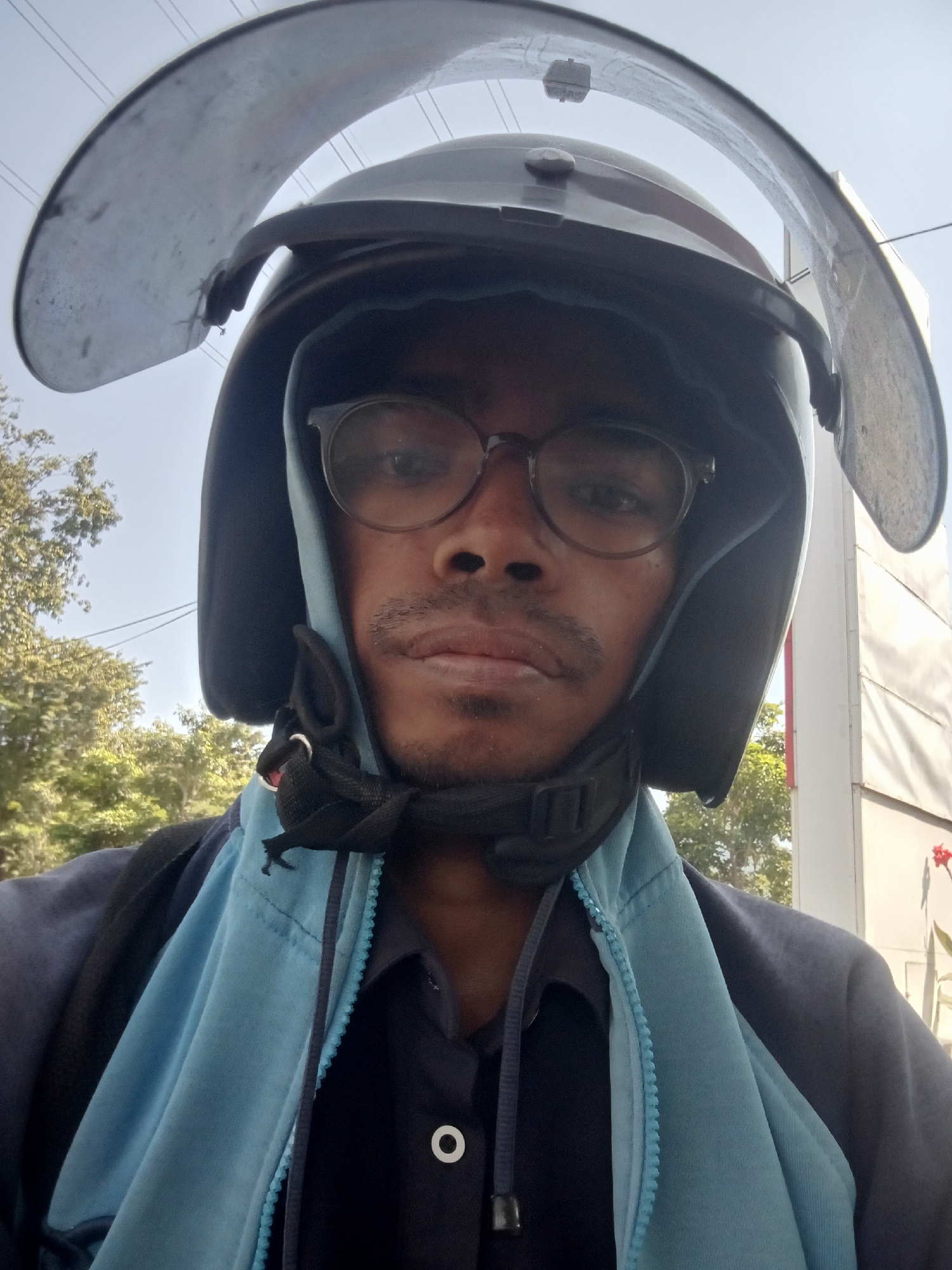 A dark-skinned person wearing a dark helmet with a clear visor and round glasses is taking a selfie outdoors. The person has a mustache and a small beard. They are wearing a light blue hooded jacket over a dark shirt with a white button visible. The background shows a bright sky, trees, and a building with a light-colored exterior. Power lines are visible in the sky. The scene is likely in an urban environment, possibly Surabaya, Indonesia, given the outdoor setting and infrastructure.