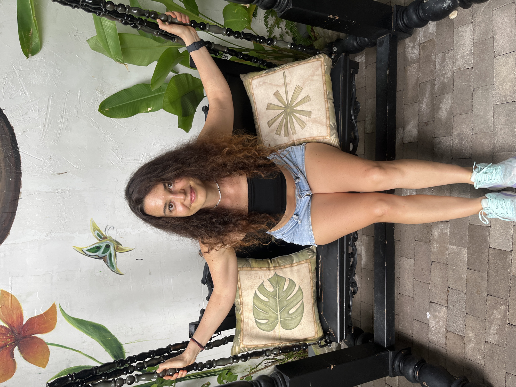 A woman with long, curly brown hair is sitting on a black swing. She is wearing a black tube top and denim shorts. She is smiling and looking at the camera. The swing has two decorative pillows on it, one with a monstera leaf design and the other with a fan-like design. Behind the swing is a white wall with a painting of an orange flower and a green butterfly. There are also large green leaves from a plant to the right of the swing. The swing is on a patio made of cobblestones. The scene appears to be in an outdoor area, possibly a garden or courtyard.