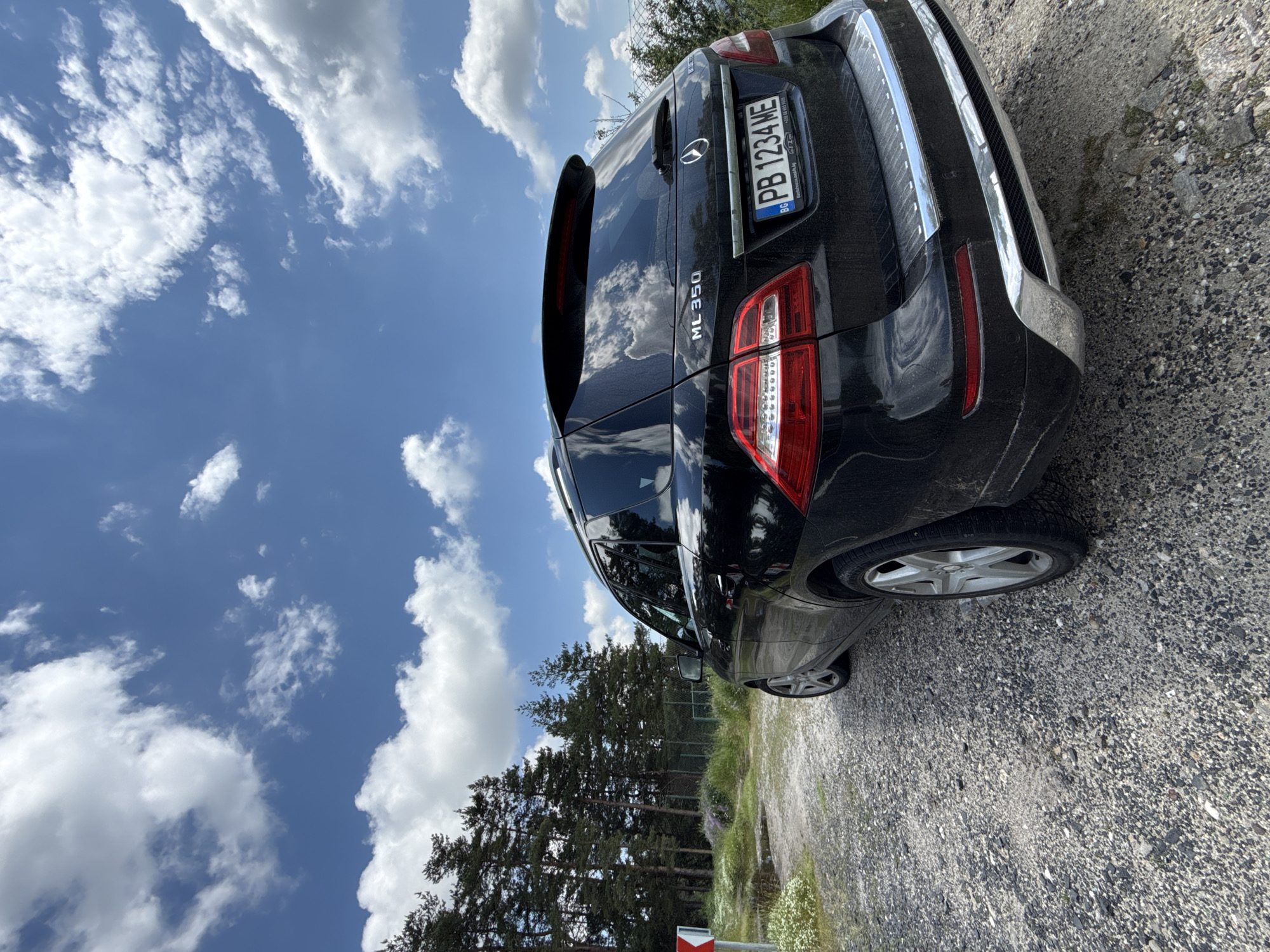 A black Mercedes-Benz ML 350 SUV, observed from its rear-right quarter, is parked on a gravel and dirt surface in the vicinity of Chubrika, Bulgaria. The vehicle bears a Bulgarian license plate, "PB 1234 ME", and shows signs of dust accumulation on its body. The model designation "ML 350" is affixed to the left side of the tailgate. Chrome trim accents the rear bumper and the lower edge of the tailgate. To the left of the vehicle, a dense cluster of tall pine trees forms a background, with green grass and small white and purple wildflowers growing at their base. A partial red and white chevron sign is visible on the far left. A fence line and sparse vegetation are discernible beyond the vehicle's right side. The sky, occupying the upper two-thirds of the frame, is clear blue with numerous scattered white cumulus clouds. The scene is illuminated by natural daylight. No people are present.