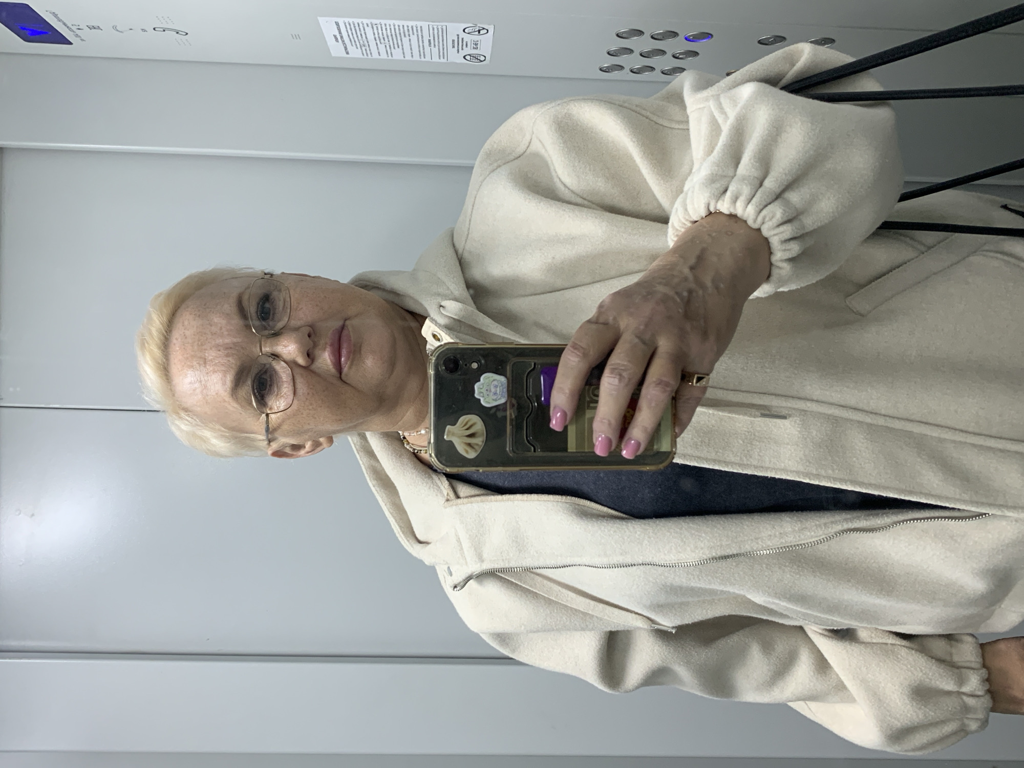 An older white woman with short blonde hair and glasses stands in an elevator, holding a cell phone in her right hand to take a selfie. She wears a beige hooded jacket over a dark grey shirt. The phone case is transparent with some small decorations. Her nails are painted pink, and she wears a ring on her right ring finger. The elevator has a grey metal interior, with panels and buttons visible. There's also a safety information sticker on the elevator wall. Based on other details and context, this image was likely taken in Chaikovsky, Russia.