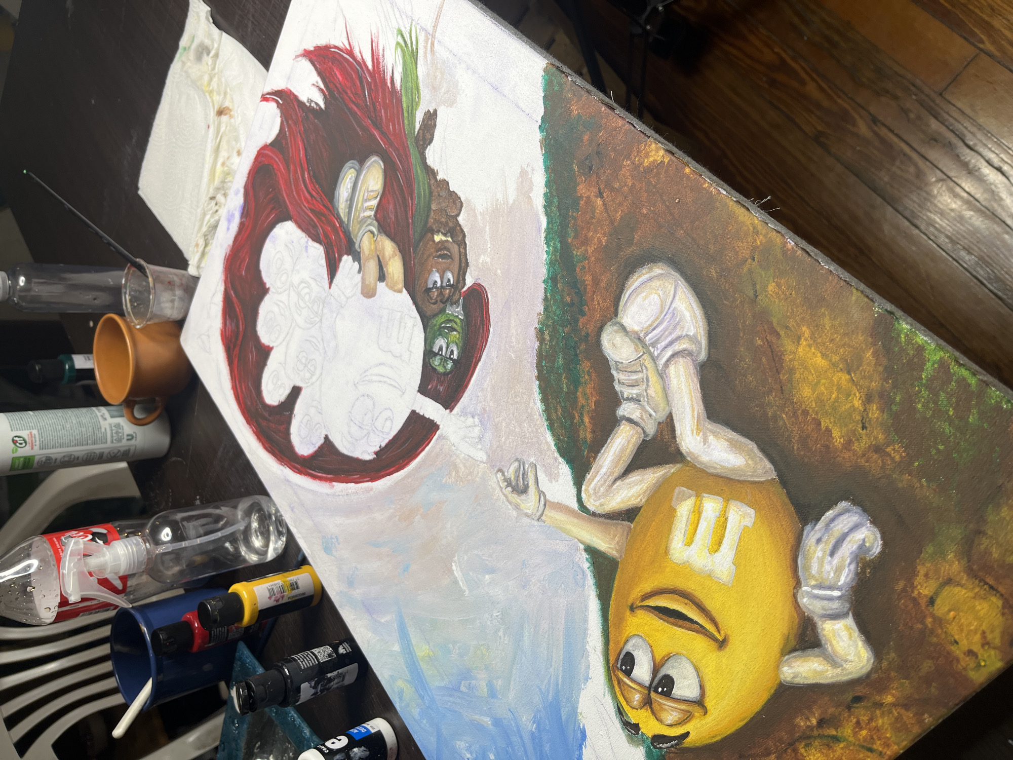 A high-angle shot displays an unfinished painting resting on a dark wooden surface. The painting presents a comedic reimagining of Michelangelo's "The Creation of Adam." On the right half of the painting, a fully rendered yellow M&M character hangs upside down, emerging from a brown and green background, its hand extended. The left half is in progress; it features an outline of white figures, including what appears to be M&M characters, one red, one brown, and one green, suspended in a red circle. The canvas is surrounded by various art supplies including paint pens, spray bottles, a glass and a mug. The location is not provided in the image.