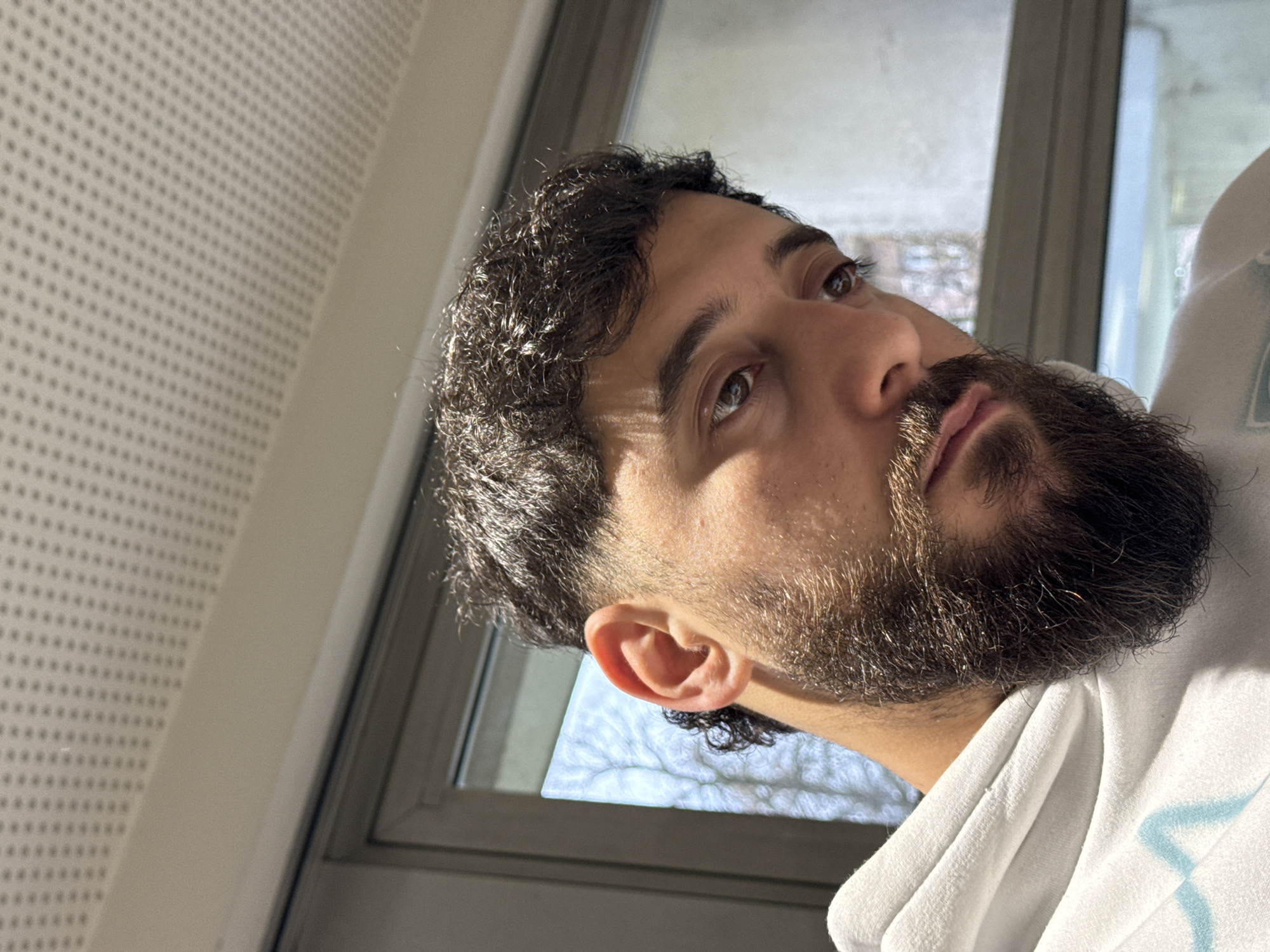 The image displays a close-up portrait of a single male subject positioned indoors. The subject has dark, curly hair with a faded haircut on the sides, a full dark beard, and brown eyes. He is wearing a white hooded sweatshirt with a light blue, wavy graphic design visible on the upper left chest area. His head is slightly tilted, and his gaze is directed downwards and to the right. The background features a perforated acoustic ceiling tile at the top of the frame, indicating an interior space. Below the ceiling, a gray-framed window is visible, through which natural daylight enters. The view outside the window shows bare tree branches against a bright sky, consistent with an urban outdoor environment. Although the location is specified as Strasbourg, France, no identifiable landmarks or specific architectural features are discernible within the image to pinpoint a precise location within the city.