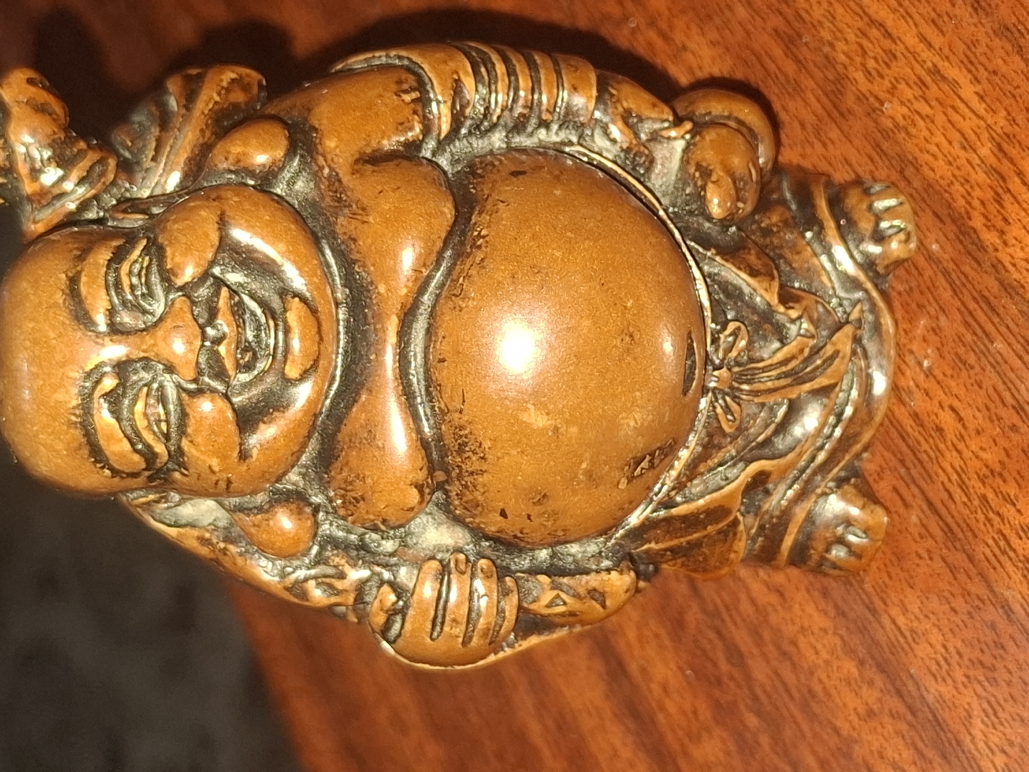 A close-up, eye-level shot shows a small, intricately carved wooden figurine of a laughing Buddha. The figurine is a warm, reddish-brown color and rests on a polished, dark wood surface, likely a table or desk. The Buddha is depicted with a prominent belly, a wide smile, closed eyes, and rosary beads around its neck. It carries a sack over its left shoulder, and its hands are clasped together. The carving details are visible, including folds in the clothing and the texture of the wood. The lighting creates highlights and shadows, emphasizing the three-dimensional nature of the figurine. The background is dark and out of focus, directing the viewer's attention to the subject. There is no information about the location in Almaty, Kazakhstan, as this detail is not depicted in the image.