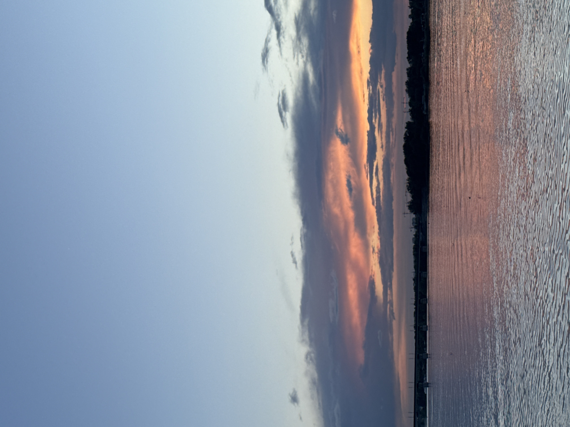 The image captures a tranquil landscape at either sunrise or sunset over a vast body of water, likely a river or large lake, within the context of Perm, Russia. The scene is bisected horizontally by a distinct horizon line.
The upper portion of the frame is dominated by an expansive sky. On the left, it transitions from a clear, pale blue to a more complex arrangement of clouds towards the right. A prominent band of dark, heavy clouds stretches across the sky, dramatically lit from behind and within by the low sun, creating vibrant splashes of orange, pink, and deep purple. Brighter orange and yellow tones are visible through gaps in these darker cloud formations, indicating the presence of the sun near the horizon.
Below the sky, a narrow, dark silhouette of land forms the horizon. This distant shoreline is densely covered with trees, and faint outlines of structures, possibly power lines or small buildings, can be discerned.
The lower half of the image is filled by the water's surface, which acts as a vast, reflective mirror. The water is covered in numerous small ripples, creating a textured pattern. It beautifully reflects the sky's colors, showing warm pink and orange hues where it mirrors the dramatic clouds, and cooler blue-grey tones where it reflects the clearer sky. A few tiny, dark specks, possibly distant birds, are visible over the water.
The overall impression is one of serene natural beauty, characteristic of dawn or dusk. There is no visible text or direct human activity captured in the frame, apart from the distant structures on the horizon.
