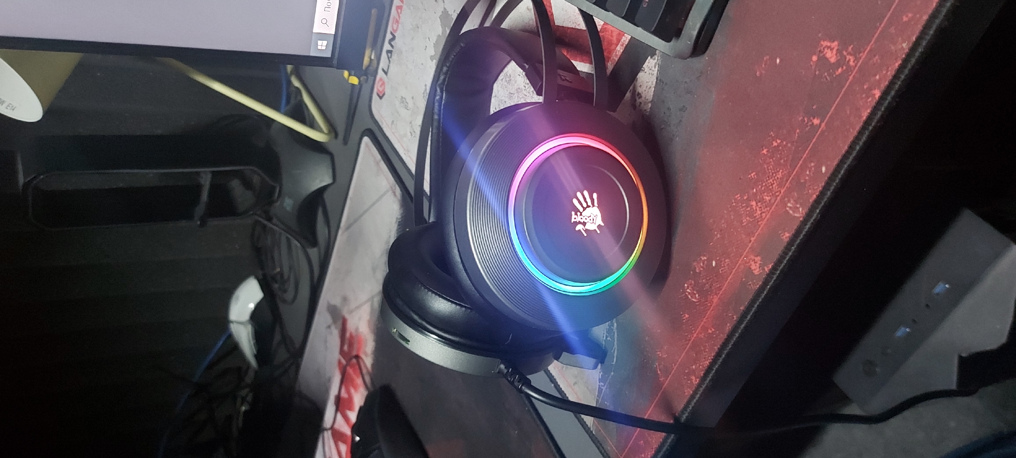 The image shows a headset resting on a mousepad. The headset is black and gray with a circular design on the earcups that features a multicolored LED ring and a white handprint logo with the word "bloody" underneath. The mousepad has a red and white design. Adjacent to the mousepad is a keyboard. Part of a computer monitor is visible. A power strip is seen with indicator lights lit. The image was taken indoors. There is no information to suggest that the image was taken in Tashkent, Uzbekistan.