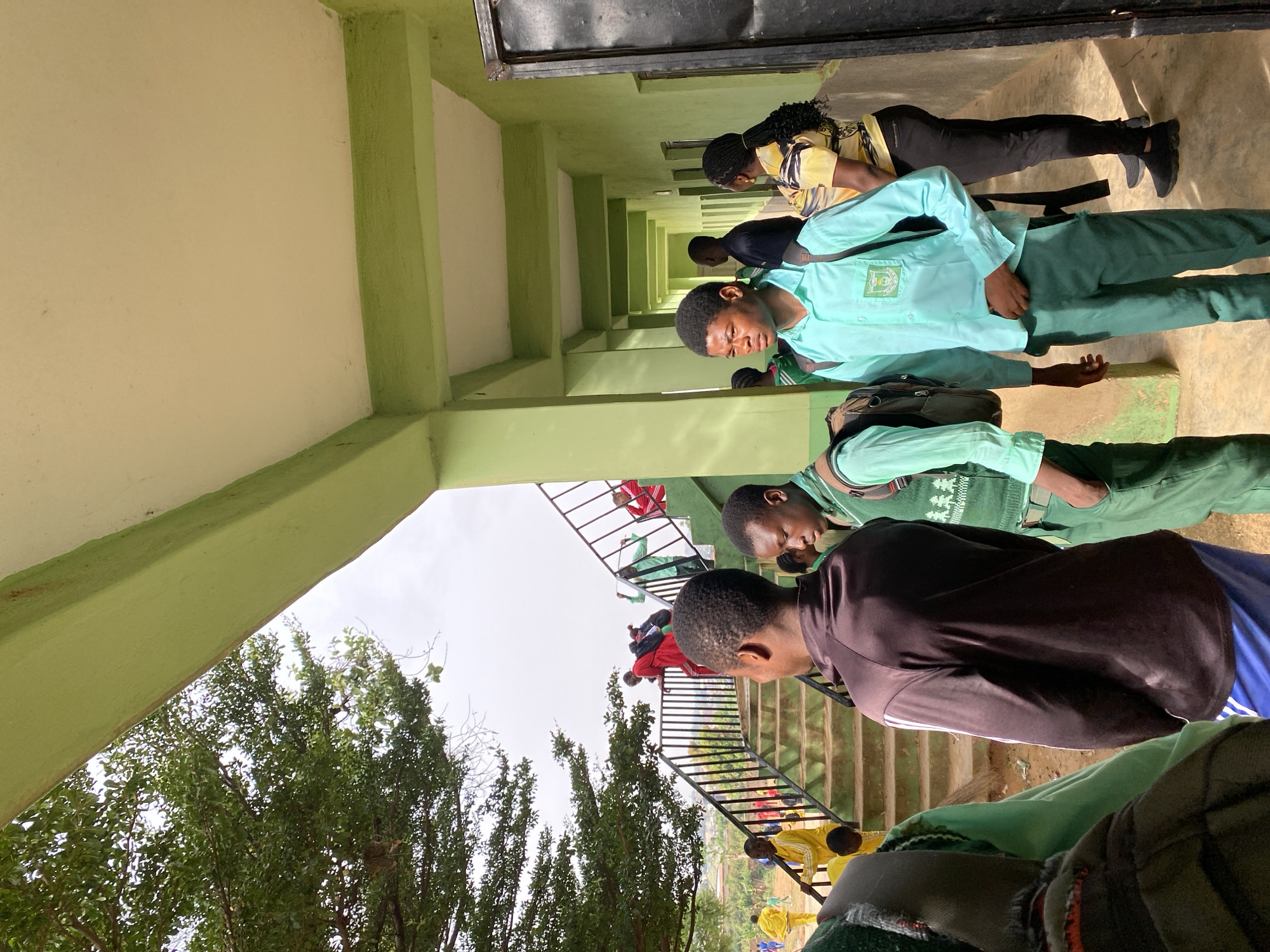 The image depicts a group of people, seemingly students, in what appears to be an outdoor corridor or walkway of a building. The structure is painted with light green and cream-colored paint. The corridor is lined with rectangular columns. Some individuals are in school uniforms, including light blue shirts and green pants. A set of stairs leads to a lower level, where other people are visible, some wearing red and yellow clothing. Trees are seen through the opening near the stairs. The location is Gura Top, Nigeria.