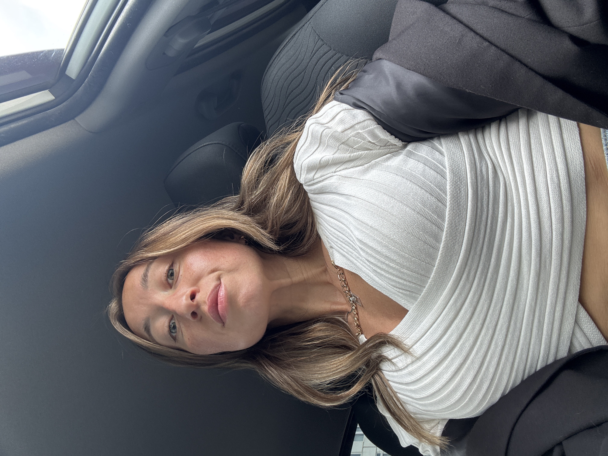 A young woman is captured in a close-up selfie inside a car, presumably in Chaikovsky, Russia. She is seen from the chest up, with her head slightly tilted and a calm, subtle smile on her face as she looks directly at the camera. She has long, light brown hair with blonde highlights, which flows over her shoulders and chest.
She is dressed in a fitted white ribbed top with long sleeves, and a dark jacket or blazer is draped over her shoulders. Around her neck, she wears a thin, gold-colored chain necklace with a small, indistinct charm.
The background reveals the interior of a car, including a dark fabric seat to her left and the car's headliner and window frame above. Bright natural light is visible entering the vehicle from above and to the left, indicating it is daytime. There is no visible text in the image.