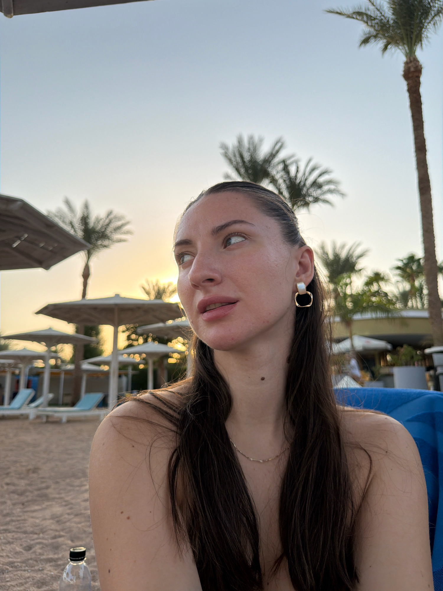A woman with long, dark hair is seated on a beach at sunset. She is looking to her right, away from the camera. She is wearing a gold earring and a delicate necklace. In the background, palm trees, beach umbrellas, and lounge chairs are visible. The sky is a pale blue, transitioning to orange near the horizon. The scene suggests a tropical resort location.