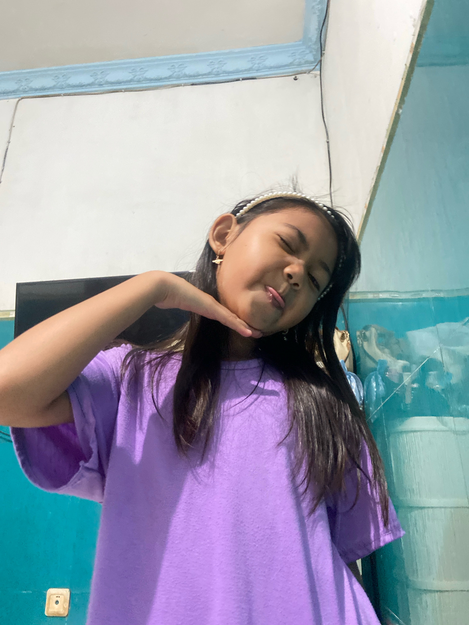 An Indonesian child, appearing to be between 6 and 10 years old, poses for a selfie in what appears to be a residential interior. The child is wearing a purple t-shirt and a pearl-beaded headband, with small gold planet-shaped earrings. Their eyes are closed, and their tongue is slightly protruding as they touch their chin with their left hand. The background features a white wall with a light blue decorative border at the top, and a turquoise wall to the right. A black television screen is visible behind the child's left shoulder. The environment suggests a domestic setting in Gelam, Indonesia.