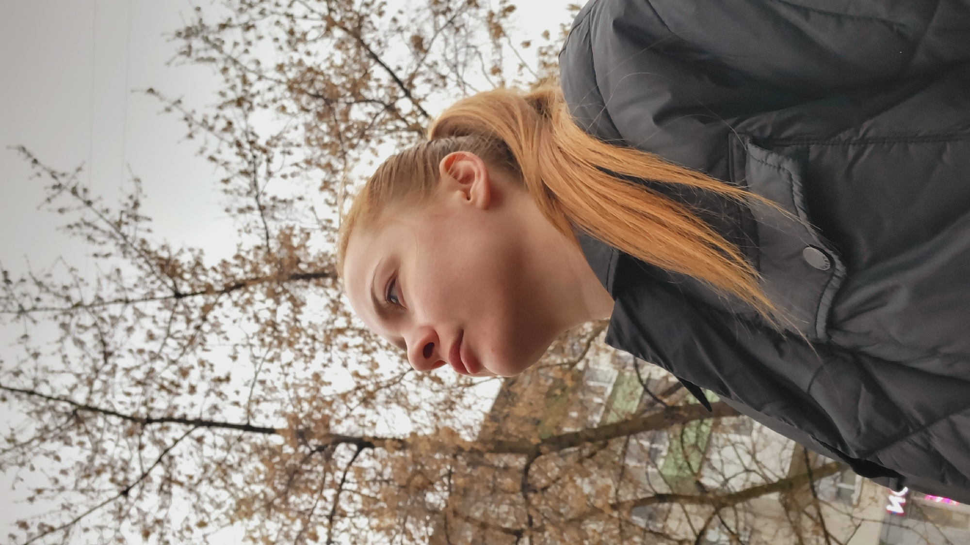 A low-angle close-up shot shows a young woman outdoors. She has fair skin, red hair pulled back into a ponytail, and is wearing a dark-colored, quilted jacket. The background includes bare branches of trees against an overcast sky and glimpses of buildings. The photo was taken in the city of Chaikovsky, Russia.