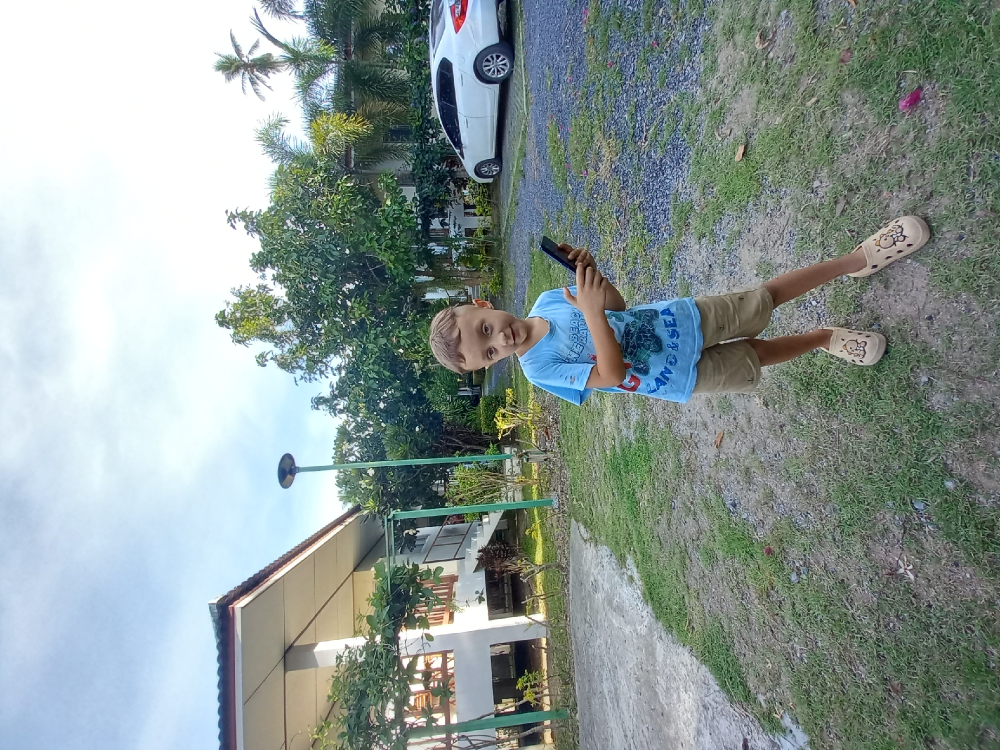 In Baan Mae Nam, Thailand, a young boy stands on a gravelly driveway with patches of grass, holding a smartphone. He is wearing a light blue t-shirt with a graphic print and tan shorts, paired with tan clogs decorated with bear imagery. The boy is positioned in front of a multi-story building with white walls and a brown-tiled roof, accessible by an outdoor staircase. To the right of the building, a white sedan is parked. The environment features lush green trees, including palm trees, and a clear sky with some clouds. A green lamp post stands near the building.
