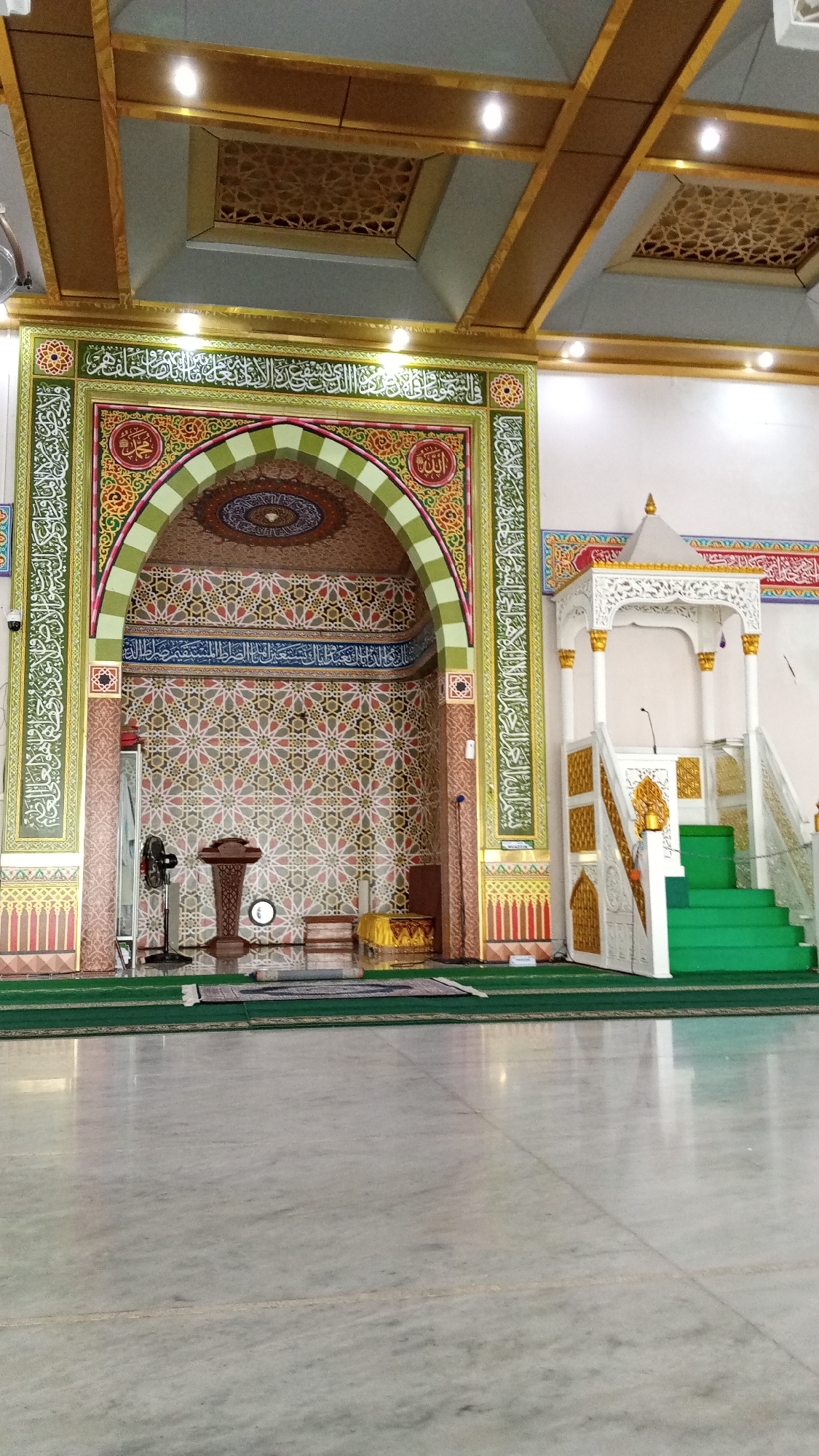 The image shows the interior of a mosque in Julikeude Dua, Indonesia. The main focus is the mihrab, an ornate alcove indicating the direction of prayer, which features intricate geometric patterns and Arabic calligraphy. To the right of the mihrab is a minbar, a pulpit from which the imam delivers sermons, decorated with white and gold accents and green carpeted steps. The ceiling is adorned with golden coffers and recessed lighting. The floor is polished marble. In front of the mihrab, a wooden lectern, a fan, and a low bench are visible. The overall atmosphere is one of solemnity and architectural beauty.