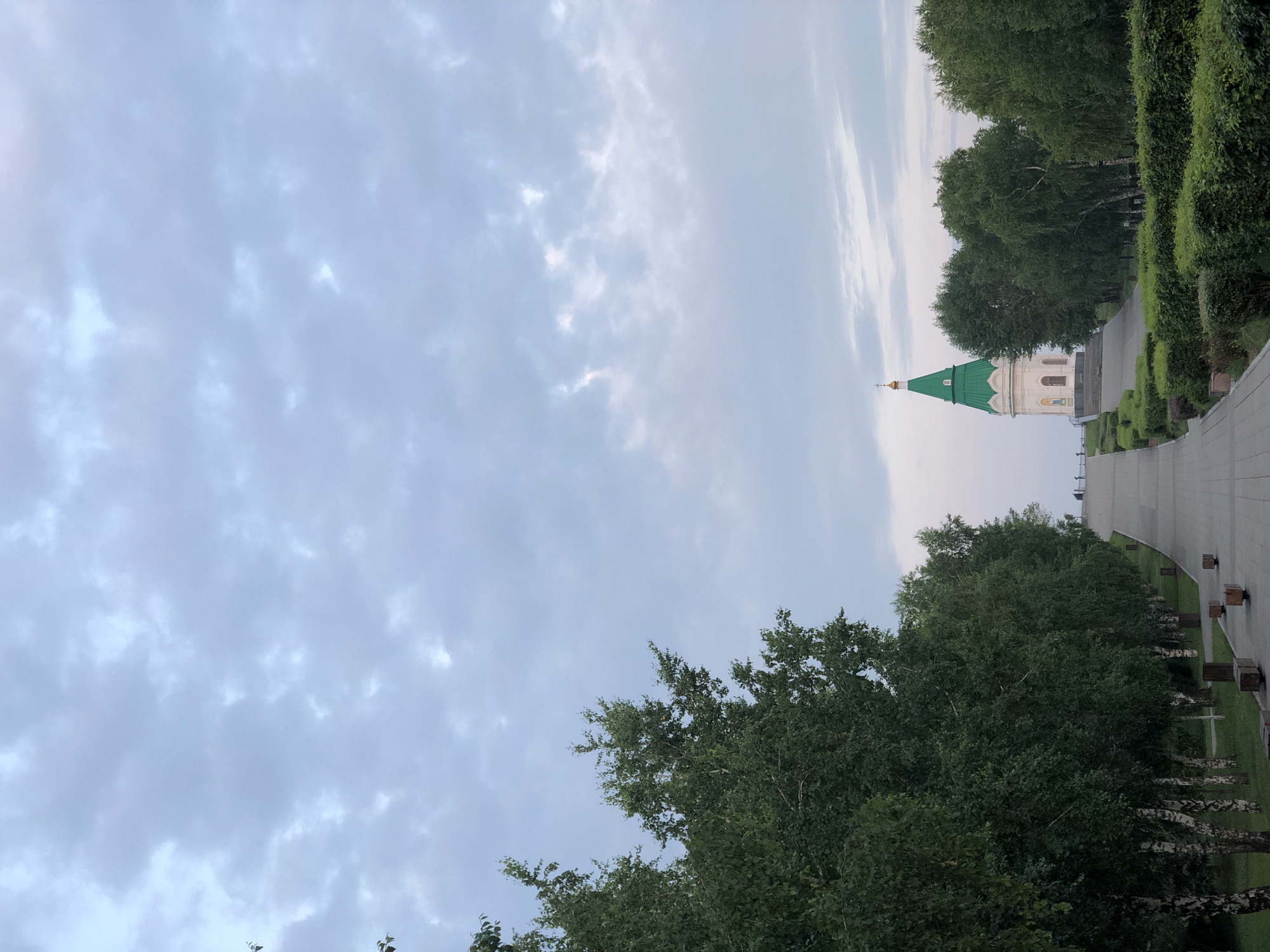 The image depicts a wide, paved pathway leading upwards towards a white building with a green, conical roof topped with a golden cross. This structure is a chapel, likely the Chapel of St. Paraskeva Pyatnitsa, a prominent landmark in Krasnoyarsk, Russia. The pathway is lined on both sides with lush green trees, creating a sense of symmetry and depth. On the left, a dense line of deciduous trees, possibly birches with their characteristic white bark, forms a wall of foliage. On the right, neatly trimmed hedges create a formal garden border, with more trees visible behind them. In the distance, at the top of the pathway, a few silhouetted figures can be seen, suggesting a public space. The sky above is overcast with a soft, diffused light, indicating either early morning or late evening. The overall atmosphere is serene and suggests a place of quiet contemplation or a popular spot for a leisurely stroll.