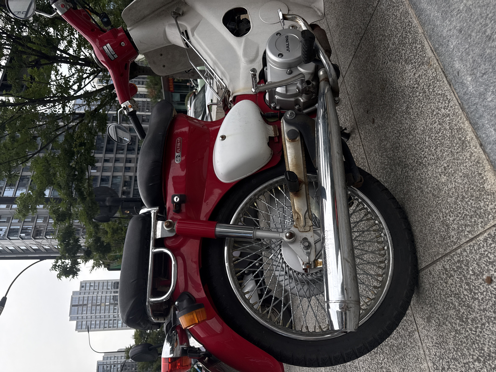 This is a medium-shot, eye-level photograph of a red and white motorcycle parked on a sidewalk in Yiwu, China. The motorcycle is viewed from the right side, with its front wheel and fender absent from the frame. The motorcycle has a red body with a black seat and a white panel on the side. The chrome exhaust pipe is visible, as well as the engine, which is branded "JIALING." The motorcycle is parked on a textured gray sidewalk, with a few small brown debris visible on the ground. In the background, there are several modern, tall buildings with glass windows, suggesting an urban environment. Trees with green foliage are also visible. The sky is overcast and gray. No people are present in the image. The overall scene depicts a parked motorcycle in a city setting.