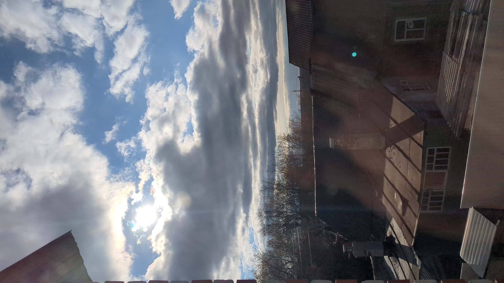 The image depicts a view from a window in Ulyanovsk, Russia. The sky occupies the left side of the frame, showing a mix of blue sky and white cumulus clouds, with the sun obscured by clouds. To the right of the clouds, a series of elongated grey clouds stretch towards the horizon. In the middle ground, bare trees are visible. On the right side of the image, part of a building is visible. The building appears to have a tiled roof and multiple windows. The building's exterior is in shades of brown and grey, suggesting it is an older structure. The bottom of the frame reveals what appears to be part of a railing or ledge. The sun glare is visible through the window, which seems to be an obstruction in the photograph.
