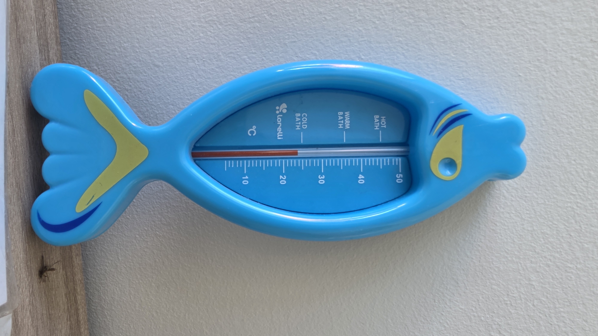 An overhead shot shows a bright blue, fish-shaped baby bath thermometer with yellow accents and a red temperature indicator. The thermometer displays measurements in degrees Celsius, with markings for "HOT BATH," "WARM BATH," and "COLD BATH." The thermometer is positioned vertically against a textured, off-white wall, with the bottom portion resting on a wooden shelf. A small, dark insect, possibly a grasshopper, is visible on the shelf to the right of the thermometer. The image was taken in Devnya, Bulgaria.