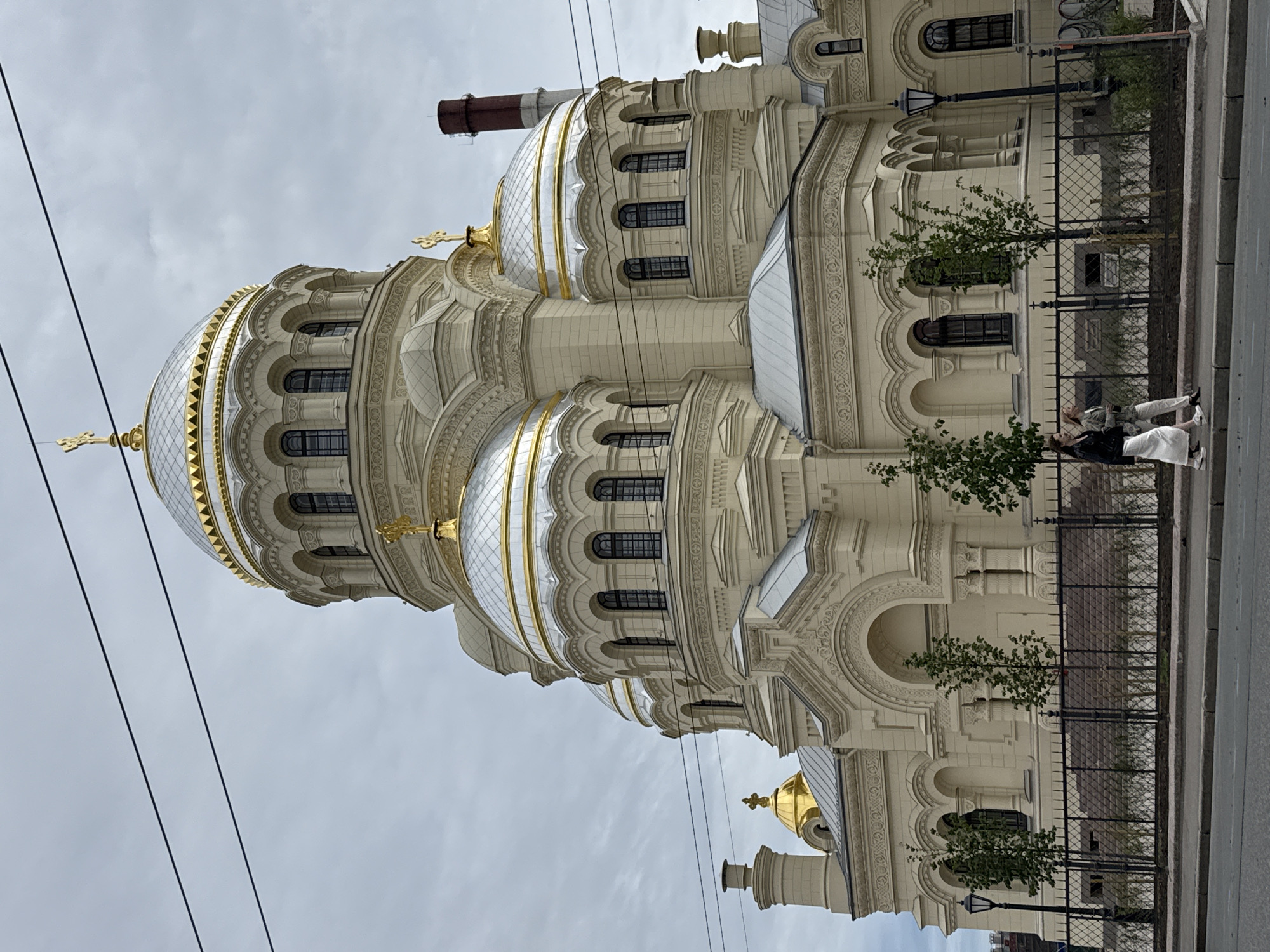 The image shows the naval cathedral of Saint Nicholas in Kronstadt, Saint Petersburg, Russia, on an overcast day. The cathedral is a large, cream-colored building with multiple domes, several of which are silver with gold accents. A prominent gold cross is visible atop the main dome. In the foreground, two women walk along a sidewalk in front of a black metal fence. The sidewalk is adjacent to a paved road. Trees are planted behind the fence. Overhead, power lines are visible against the cloudy sky. A reddish-brown chimney stack is also visible to the right of the cathedral.