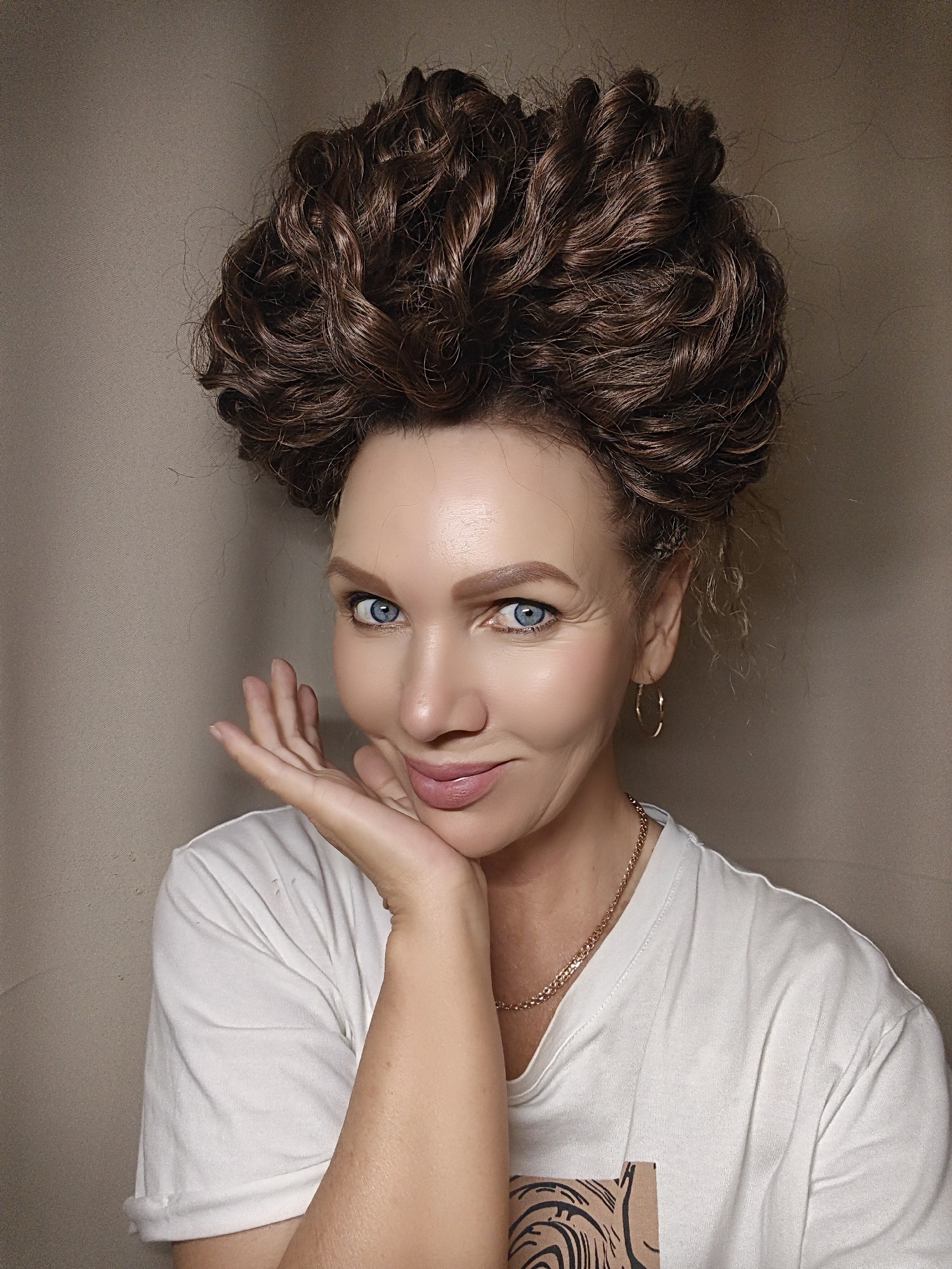 The image is a medium-shot portrait of an adult female with fair skin, pronounced blue eyes, and dark brown hair styled in a high, voluminous, tightly-curled updo. Her face displays a subtle smile with pink-toned lipstick. Her eyebrows are well-defined. She wears a plain white crew-neck t-shirt, the left sleeve of which is rolled up. A faint, abstract, brown-and-black graphic design is partially visible on the lower right front of the shirt. Accessories include a thin gold chain necklace and a single gold hoop earring visible on her right ear. Her left hand, with fingers slightly splayed, is positioned beneath her chin and jawline, serving as a prop. The background consists of a solid, light beige or tan wall, indicating an indoor setting. There are no additional objects or discernible features in the background. The lighting is soft and even, illuminating her face and hair. The location is an unspecified indoor setting in the city of Unknown, Unknown.