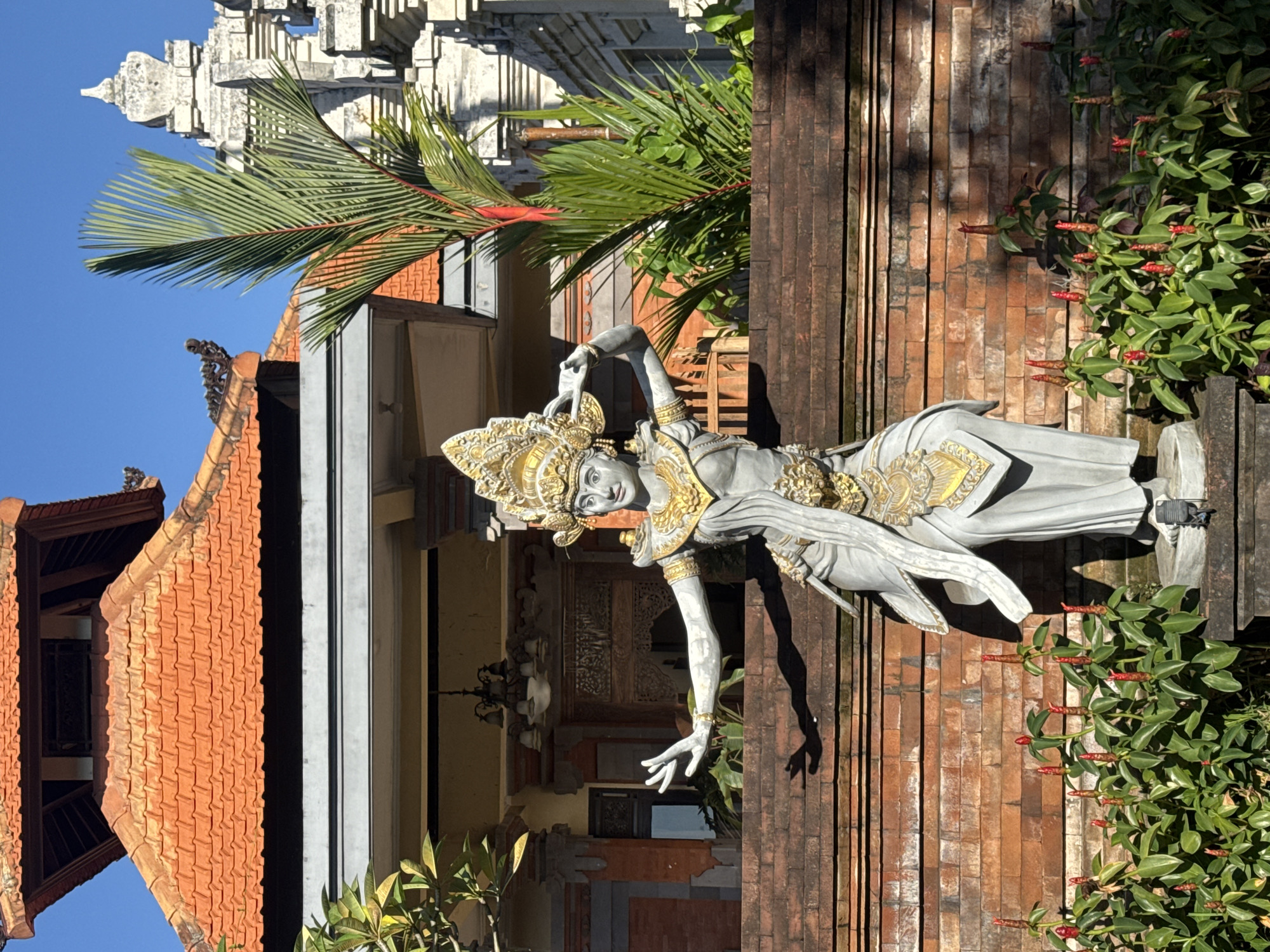 The image captures a Balinese statue adorned with gold detailing, positioned next to a brick wall and verdant greenery with red flowers. The architecture in the background suggests a traditional Balinese temple setting, likely located in Kutuh, Indonesia. Part of the pagoda is seen, along with a palm tree. The statue itself appears to be in a classical dance pose, with one arm extended. A building and roof are visible.