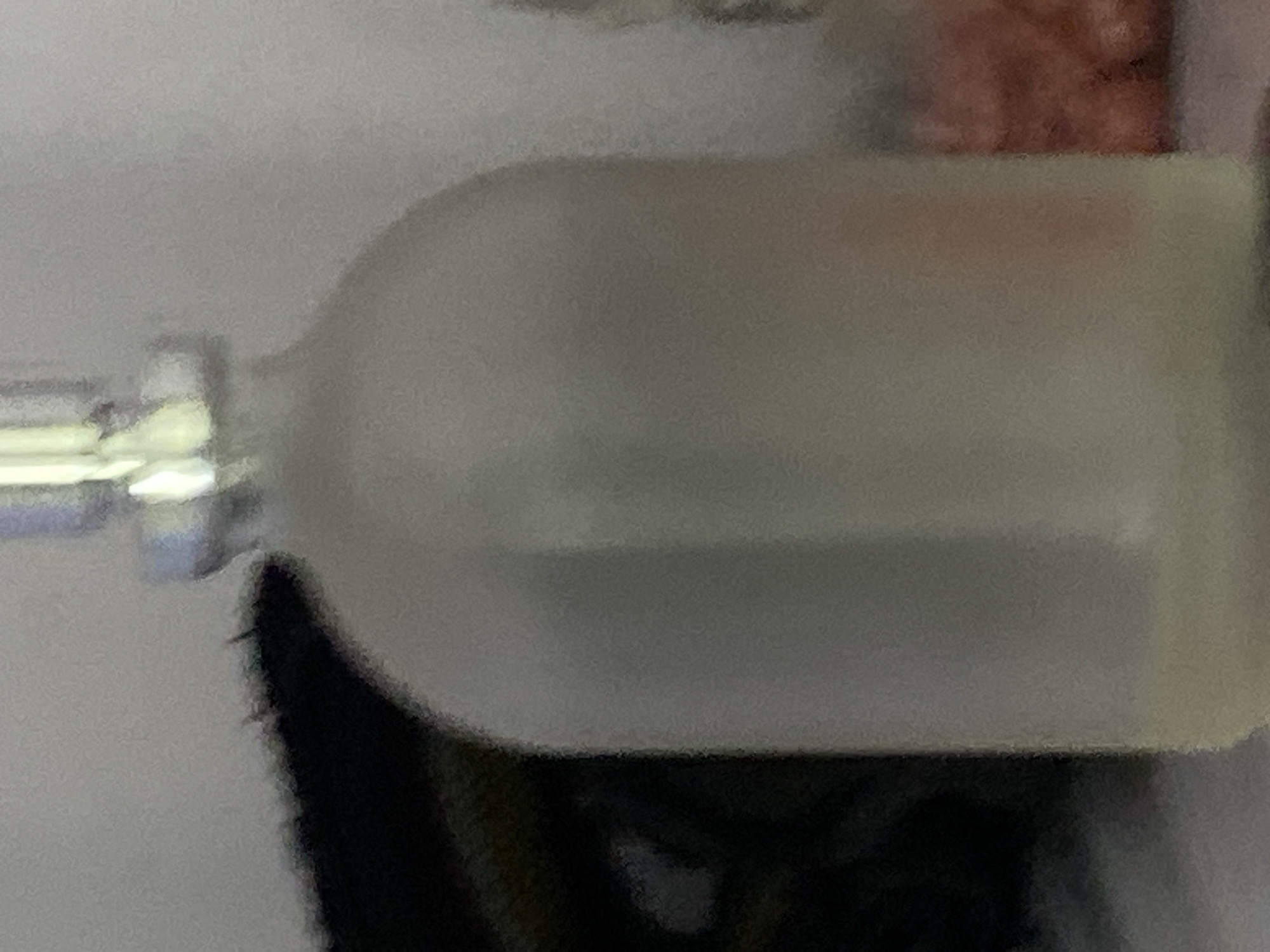 A frosted glass bottle with a silver cap stands in the foreground. The bottle is partially filled with a pale yellow liquid. To the left of the bottle, a dark, fuzzy object with sharp edges is visible. To the right of the bottle, a blurred reddish-orange object, possibly food items, is seen. The background is out of focus and appears to be a wall or similar surface. The scene is lit by artificial light, casting soft shadows. There are no people or animals present. The image does not provide any information about the location in the city of Jauro Yinu, Nigeria.