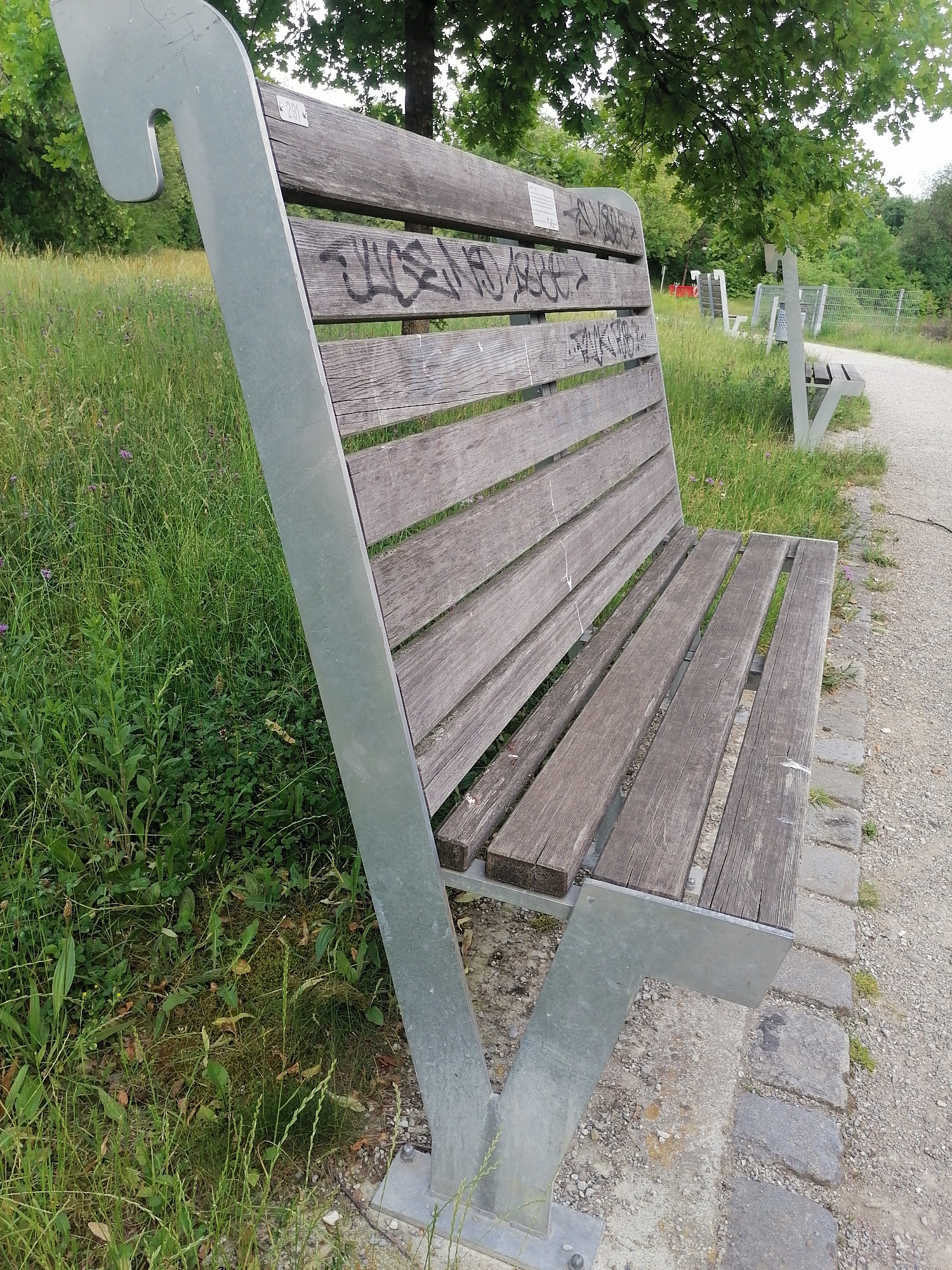 The image shows a wooden park bench with metal supports in Unterhaching, Germany. The bench is situated on a gravel path next to a grassy area with trees. The sun appears to be shining, casting subtle shadows. The bench has graffiti on its backrest, which appears to be in black spray paint. The graffiti is illegible due to its abstract nature and the image quality. In the background, other park benches and a fence are visible, suggesting a public park or recreational area. The overall scene is peaceful and natural, typical of a park setting. There are no people visible in the image. The time of day is likely daytime, given the lighting. The weather appears to be fair, with green foliage indicating a temperate season. The location cues, such as the trees and the style of the bench, are consistent with a German park.