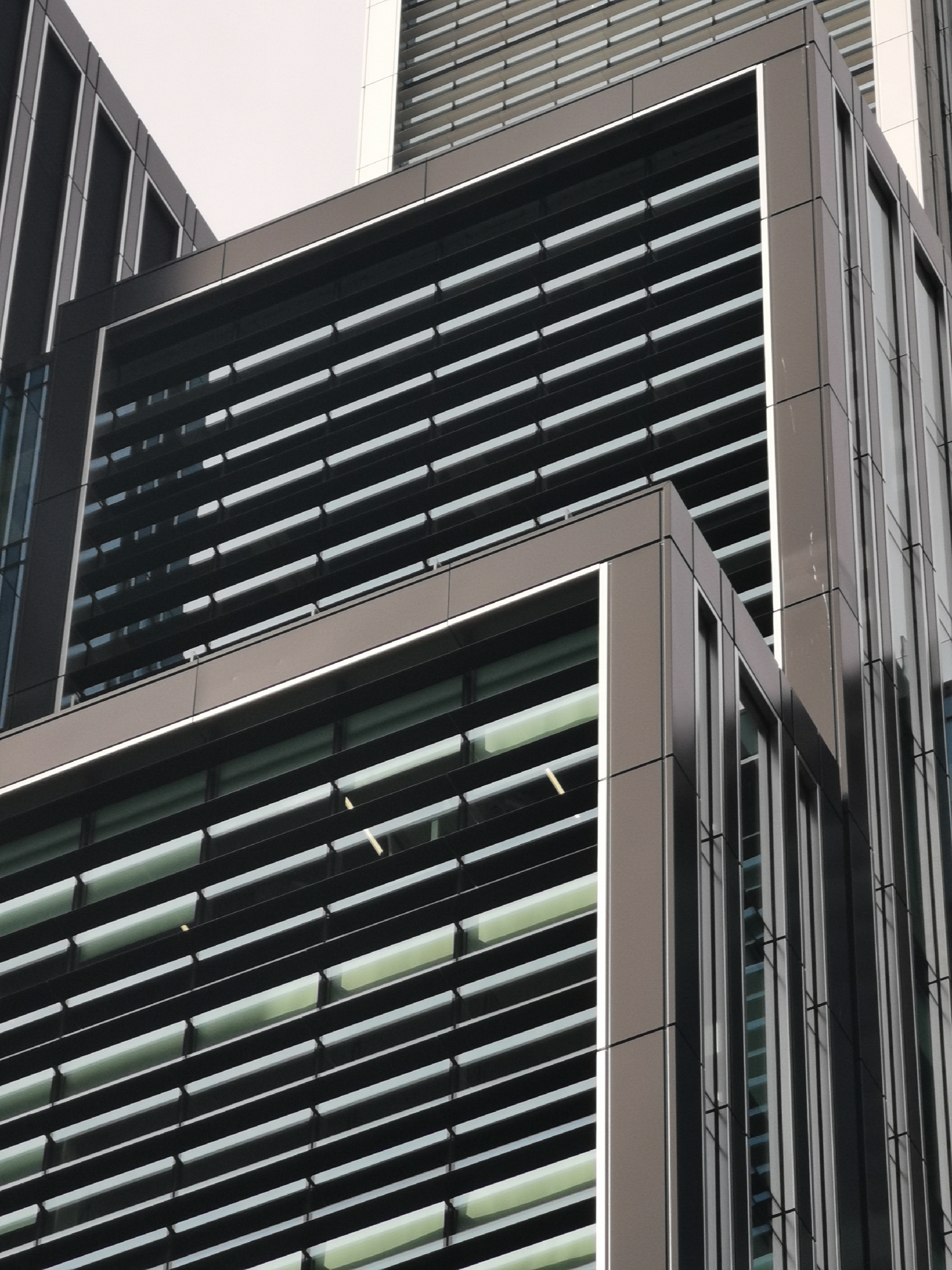 The image captures a close-up, upward-angled view of a modern building facade. The primary subject is the building's architecture, characterized by dark, possibly metallic or composite, panels and numerous horizontal louvers that cover large glass windows. The design is geometric, featuring sharp angles and defined structural lines.
No people or discernible activities are present in the scene. The setting appears to be an urban environment, but specific location cues are absent.
Notable details include the building's dark gray or black cladding, contrasting with lighter gray or white trim around the window sections and the bright, uniform white sky, suggesting an overcast day. The windows behind the horizontal louvers show hints of a greenish-yellow light, possibly from internal lighting or reflections. The perspective emphasizes the height and architectural patterns of the structure. The lighting is diffused and even, without harsh shadows, consistent with an overcast sky. There is no visible text in the image.
