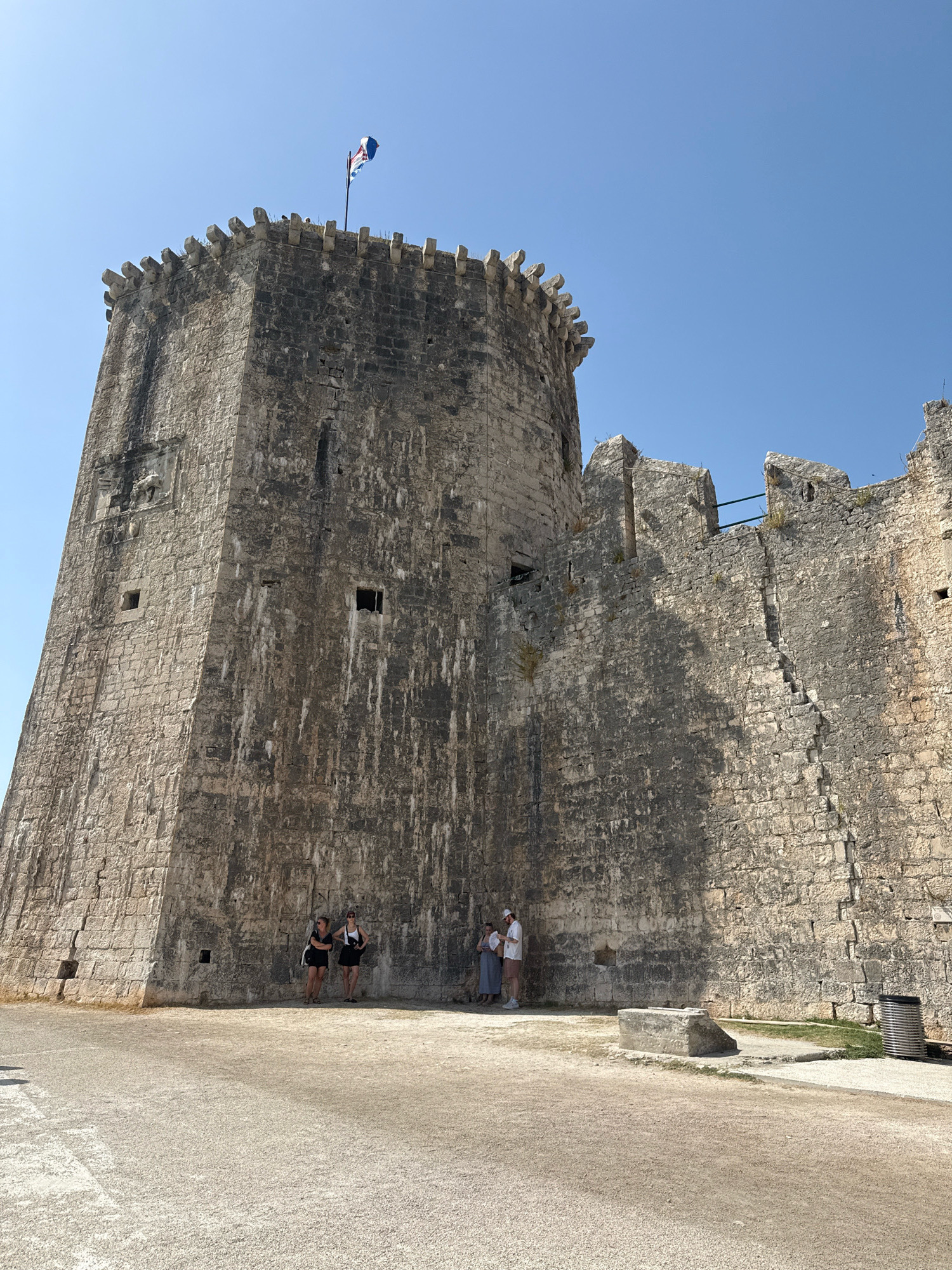 The image depicts the Kamerlengo Castle in Trogir, Croatia. It is a fortified structure built of stone blocks. The castle has a prominent round tower on the left side of the image, topped with crenellations and a flag flying from a pole. Adjacent to the tower, a section of the castle wall extends to the right, also featuring crenellations. The wall is weathered and shows signs of age, with some darker patches and cracks.
In front of the castle wall, on a gravelly ground surface, are four individuals. Two women in dark clothing are standing to the left, facing the camera. To their right, a couple, a man in a white shirt and beige shorts, and a woman in a light-colored dress, are standing and appear to be interacting or looking at something. Further to the right, a silver cylindrical trash receptacle is visible near the wall. The sky is clear and blue. The overall scene suggests a sunny day at a historical site.