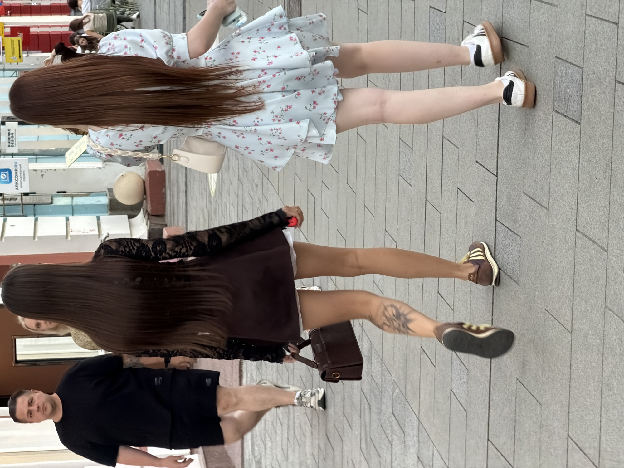 The image shows two young women walking on a sidewalk in Novaya Usman', Russia. The woman on the left has long dark hair and is wearing a dark lace top and a burgundy skirt with white shorts underneath. She is carrying a dark brown handbag. She has a tattoo on her left calf. The woman on the right has long brown hair and is wearing a light blue floral dress. She is carrying a light-colored handbag and a phone or wallet. In the background, a man in a black t-shirt and shorts is walking away from the camera. Other people are visible in the distance, as well as signs and buildings, suggesting an urban environment. The sidewalk is paved with rectangular stone tiles.