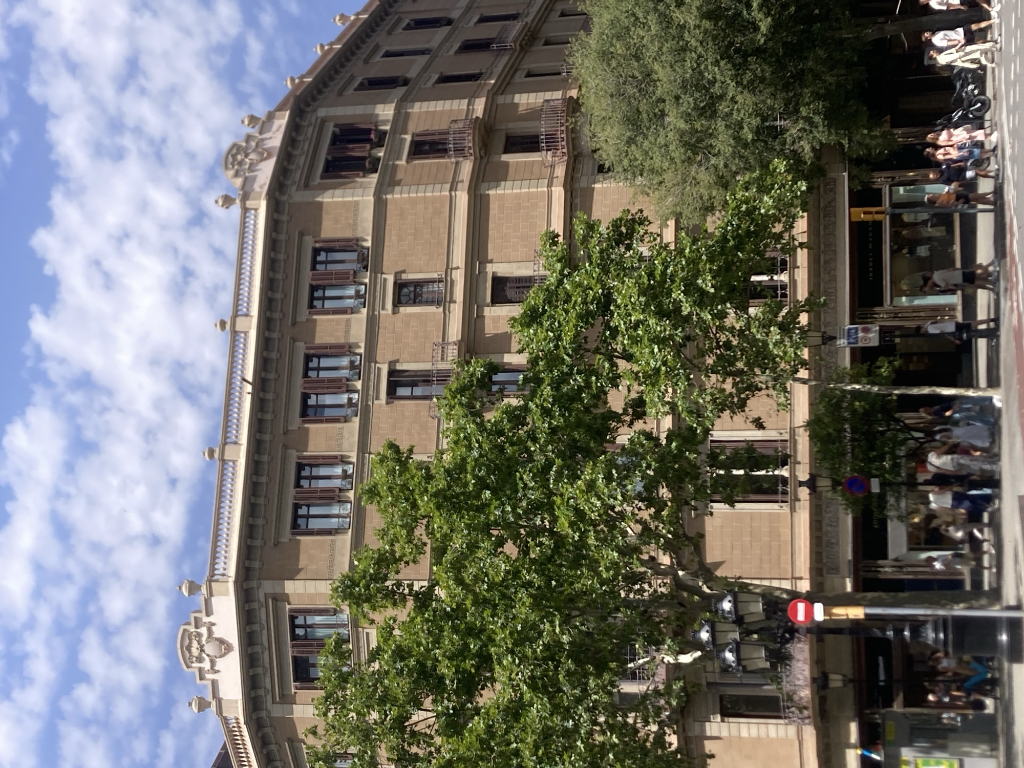 The image depicts a sunny day in Barcelona, Spain, with a view of a multi-story building and a street scene. The building features a light brown facade with numerous windows, many with dark brown shutters, and decorative balconies. The upper part of the building has an ornate cornice and rooftop detail. Lush green trees partially obscure the lower floors of the building, with their branches extending into the foreground.
The street level shows shopfronts, including one with the name "ISABEL MARANT" visible. Several people are walking on the sidewalk and crossing the street. They are casually dressed, suggesting a warm climate. Objects visible include streetlights, traffic signals, and a parked motorcycle. The sky is blue with scattered white clouds. The scene appears to be a busy urban environment, likely a commercial or residential area within Barcelona.