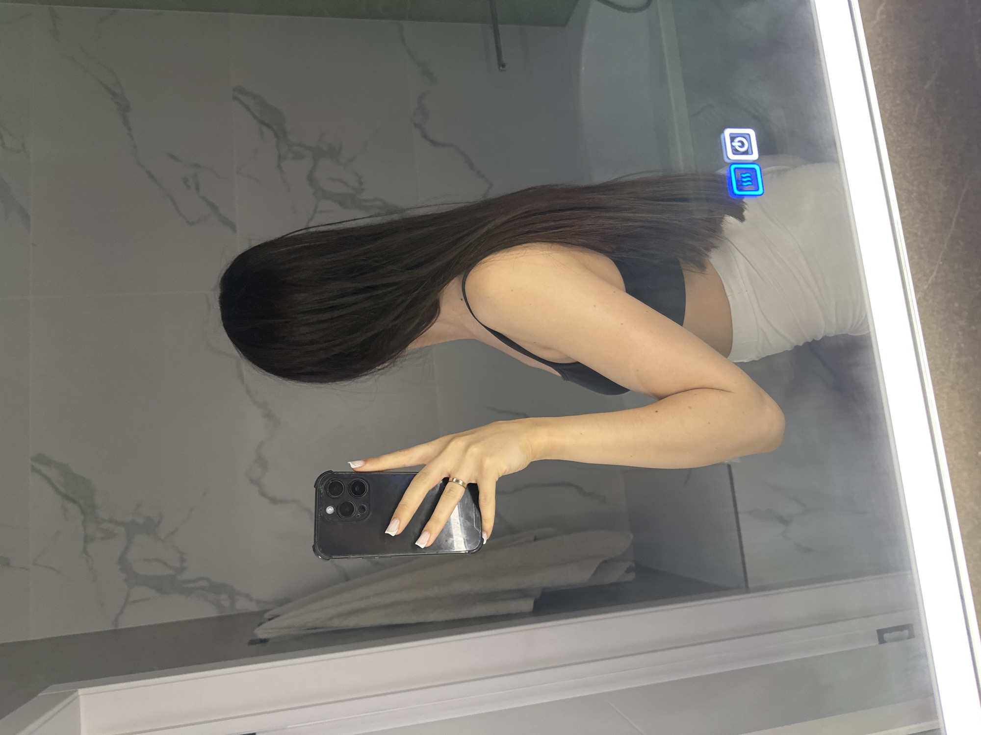 A person is taking a mirror selfie in a bathroom. The person is standing with their back to the camera, wearing a black tank top and a light-colored skirt. They are holding a black smartphone in their right hand, taking the photo. The mirror reflects the person and the bathroom environment, which includes a marble-tiled wall, a towel hanging on a rack, a bathtub, and a smart mirror with illuminated icons. The image does not provide any information about the location in the city of Estachi, Russia.