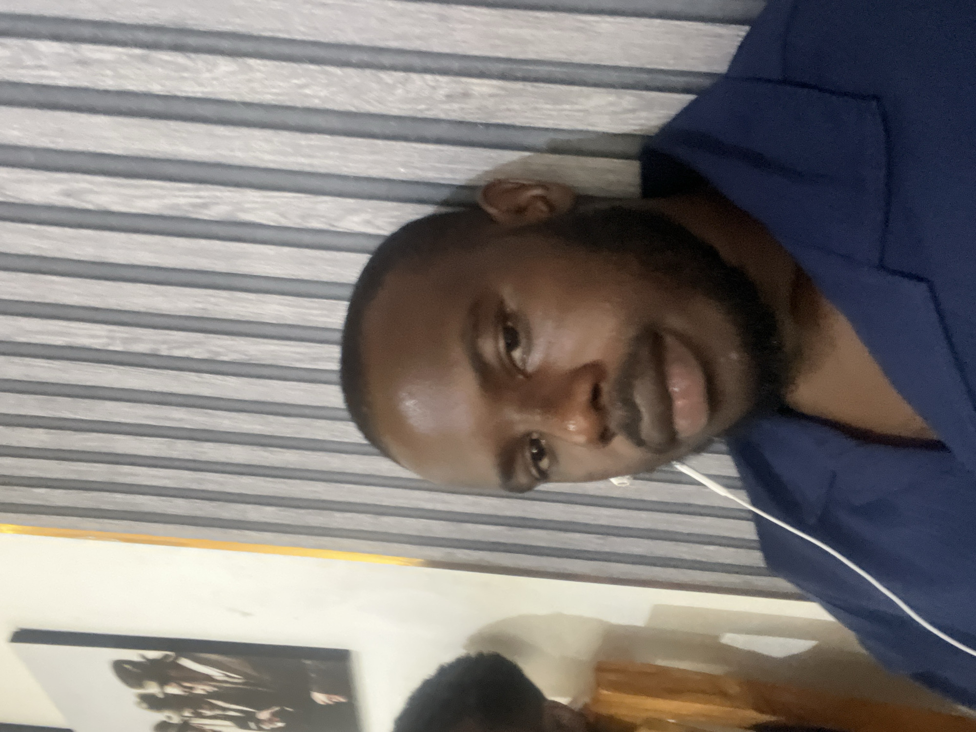 A medium shot, low-angle, slightly canted close-up of a Black man with a shaved head and a neatly trimmed beard, wearing a navy blue collared shirt, possibly a jacket or blazer. The background is a wall with horizontal, grey and white striped paneling. An earbud wire is partially visible near his neck. Beneath, near the bottom of the image is a painting on the wall, and a person, that appears to be on a couch.
There is no verifiable information to place this image in Jos, Nigeria.