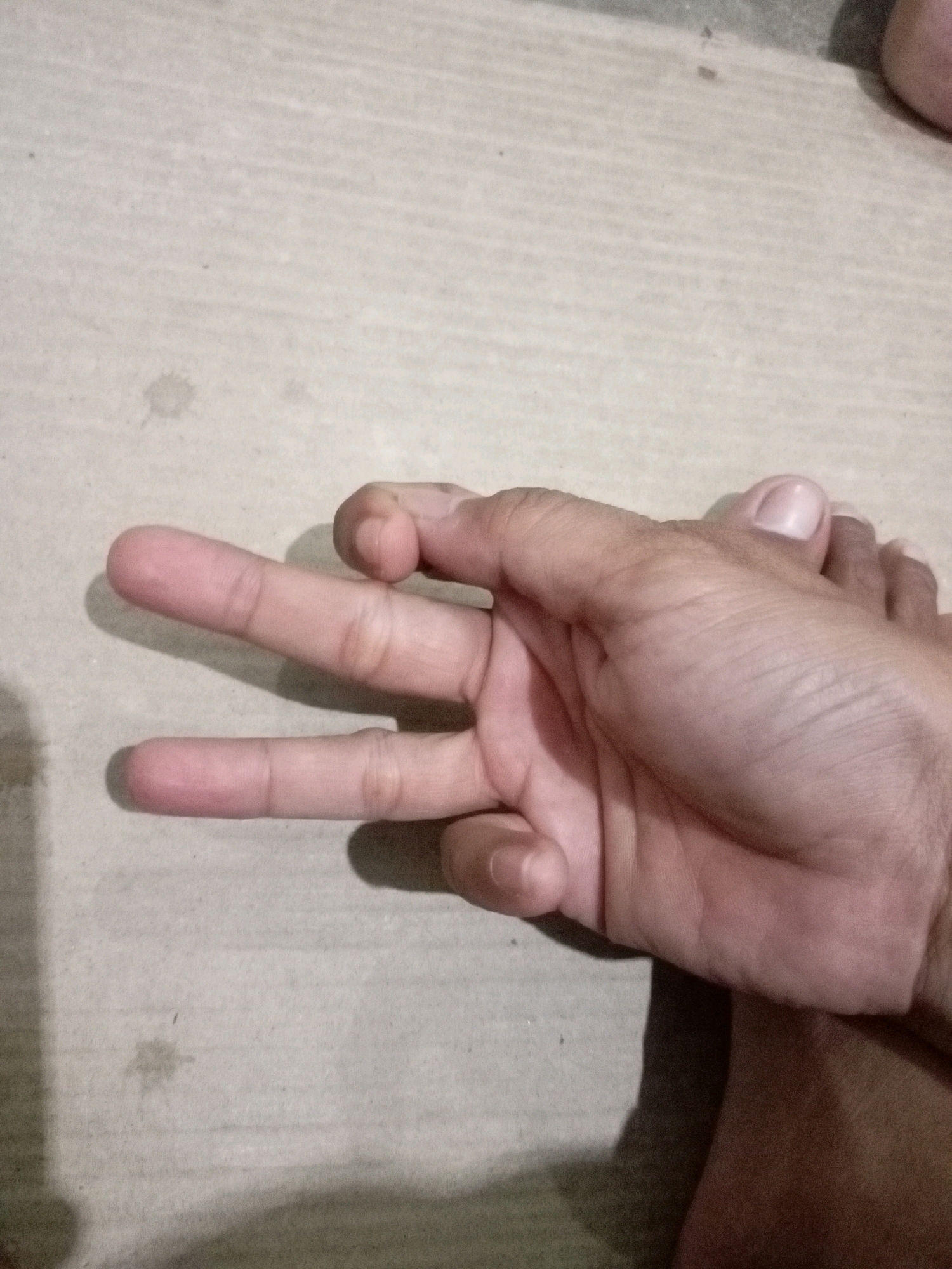 A close-up overhead shot shows a person's left hand on a light-colored textured surface, possibly concrete or a similar material. The hand is positioned with the palm facing down, and the index and middle fingers are extended upwards and separated, forming a "V" shape. The ring finger and pinky finger are curled downwards, and the thumb is bent inwards, touching the palm. The skin tone of the hand appears light to medium. A shadow is cast to the left of the hand. The background consists of the same textured surface, with subtle variations in shading and some dark specks. The image does not provide any specific information about the city of Batam, Indonesia, or any identifiable landmarks or context that would place it in that location.