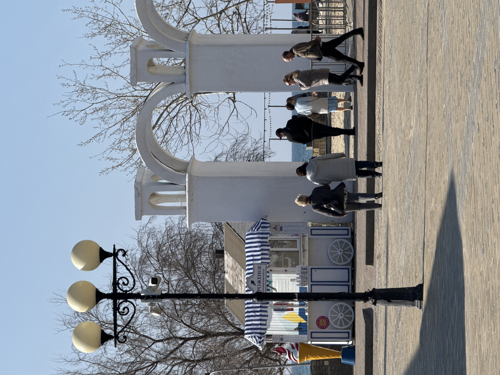 The image depicts a daytime scene on a promenade or waterfront area in Anapa, Russia, under a clear, pale blue sky.
In the immediate foreground, a black, ornate street lamp post with three cream-colored spherical luminaires stands prominently on the left. Two white security cameras are mounted on the lamp post, positioned below the luminaires. The ground is paved with light brown rectangular blocks, and a distinct shadow from the lamp post extends diagonally towards the lower left.
To the left of the lamp post and slightly behind it, a white wooden kiosk with a blue and white striped awning is visible. The word "МОРОЖЕНОЕ" (Ice Cream) is printed on the awning. The kiosk's base features decorative white circular elements resembling wheels. A large, yellow ice cream cone statue with a red and white swirl stands on a blue base to the far left of the kiosk. Bare deciduous trees are visible behind the kiosk.
A white architectural structure composed of multiple arches serves as a gateway in the midground, framing the view towards the sea. Five adult individuals are present in the central area. Two women, one in a dark gray jacket and the other in a light gray long coat, walk away from the viewer, towards the arched structure. Three other individuals are positioned under or near the arches: a man in a black jacket and baseball cap faces left; a woman in a light blue coat and dress stands facing left; and a woman in a beige coat with sunglasses walks right, appearing to converse with a man in a brown jacket who is also walking right. These individuals are on a paved path that appears slightly elevated above a sandy area.
Beyond the arches and a metal fence/railing, the calm, blue waters of the Black Sea are visible beneath the clear sky. A sandy beach stretches in front of the sea, with a string of decorative lights or banners suspended horizontally above it. Several indistinct figures are visible further down the beach. Additional bare trees with slender branches are present in the background, behind the arched structure and extending towards the horizon.