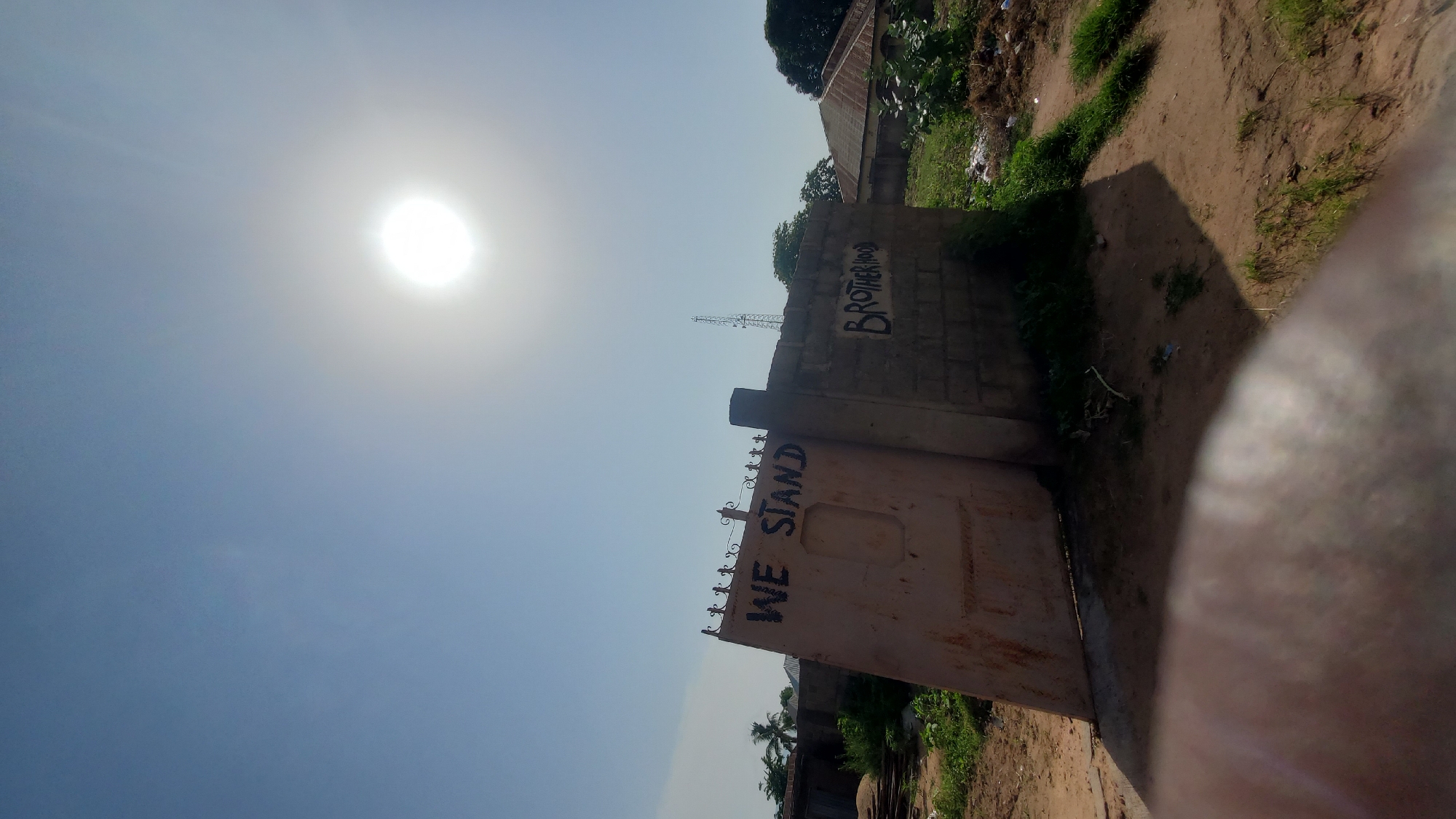 The image depicts an outdoor scene under a bright, hazy sun. In the foreground, a large, rusty metal gate is partially open, with "WE STAND" spray-painted in black letters across its surface. To the right of the gate, a brick wall has "BROTHERHOOD" spray-painted on it, also in black. The environment appears to be somewhat unkempt, with dry dirt, patches of grass, and some debris visible. In the background, buildings with corrugated metal roofs and a telecommunications tower can be seen. The overall impression is of a slightly dilapidated urban or semi-urban setting. The image appears to have been taken in Jalingo, Nigeria, as per the prompt.