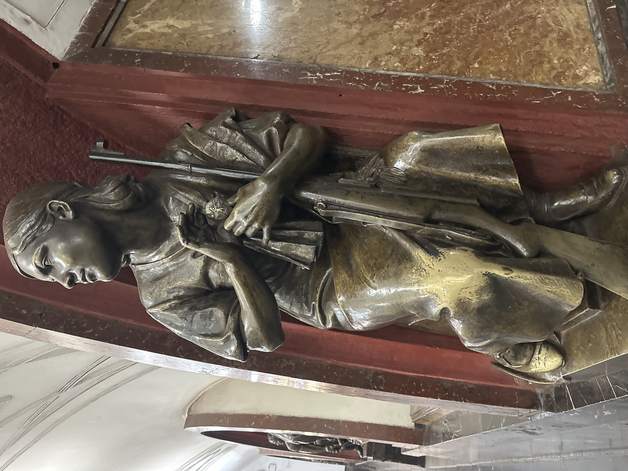 This is a medium-shot, eye-level, detailed photograph of a bronze sculpture of a woman in what appears to be a metro station in Moscow, Russia. The sculpture is affixed to a red, pillar-like structure, and a marble-patterned wall is visible to the right. The woman is depicted in a seated or kneeling pose, looking to her left with her hands clasped near her chest. She is wearing a loose-fitting garment and boots. She is holding a rifle vertically, with the butt of the rifle resting on the base and the barrel extending upwards past her head. A medal is visible on her chest, attached to her clothing. The background shows arched openings, one of which reveals another similar sculpture. The overall lighting is diffused, highlighting the metallic sheen of the sculpture.