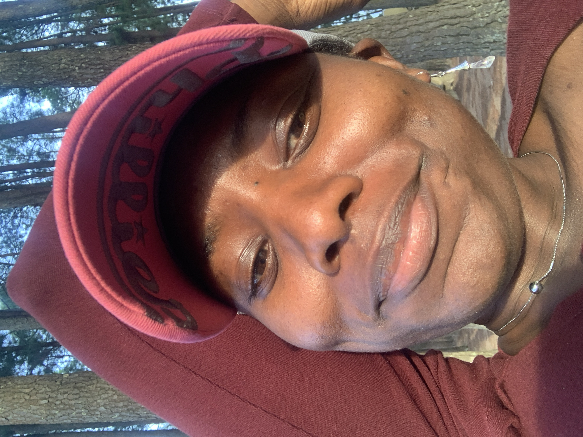 A person is captured in a close-up outdoor portrait, tilted slightly as if lying down or leaning back. The individual has a dark skin tone and is looking towards the camera with a subtle smile. They are wearing a maroon-red long-sleeved top and a matching red baseball cap. A thin silver necklace with a small spherical pendant is visible around their neck. The brim of the cap casts a shadow over part of their face.
The background consists of several tall tree trunks and sparse foliage, indicating a forest or wooded area. The bright sunlight suggests it is daytime and the weather is sunny, creating highlights on the person's skin and casting shadows. On the inside brim of the red cap, stylized dark lettering is visible, appearing to spell out "PREP" or a similar word. The overall mood is relaxed and pleasant.