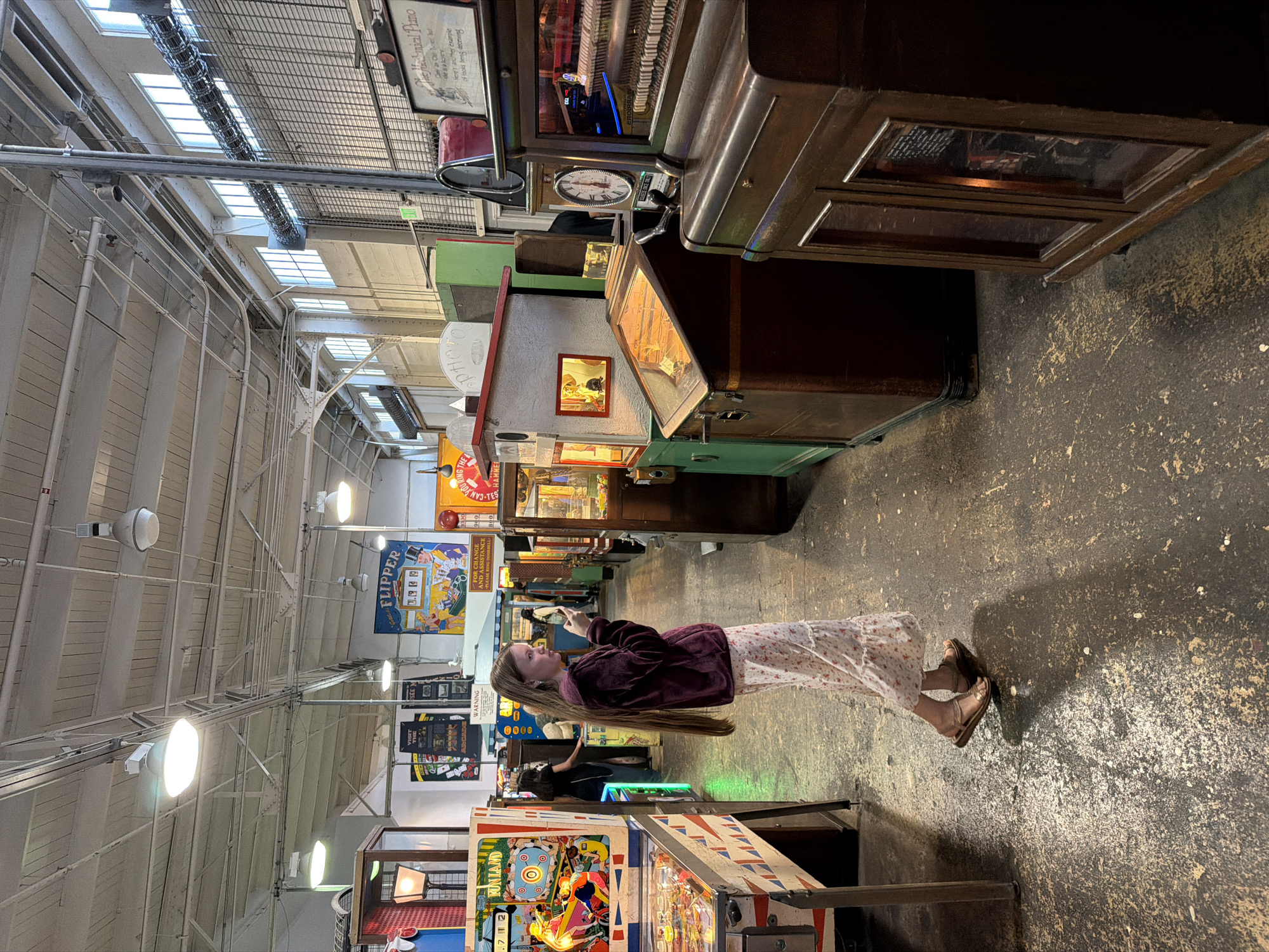 The image shows a young girl with long brown hair standing in an arcade hall in the Musée Mécanique, located at Fisherman's Wharf in San Francisco, California. She is wearing a maroon hoodie and a long floral print skirt, with her feet in sandals. The girl is holding a smartphone and looking at it. The arcade is filled with vintage mechanical games and pinball machines. In the background, other people are visible, as well as signs and displays related to the exhibits. The hall has a high ceiling with exposed pipes and lighting fixtures. The floor is made of concrete and appears worn.