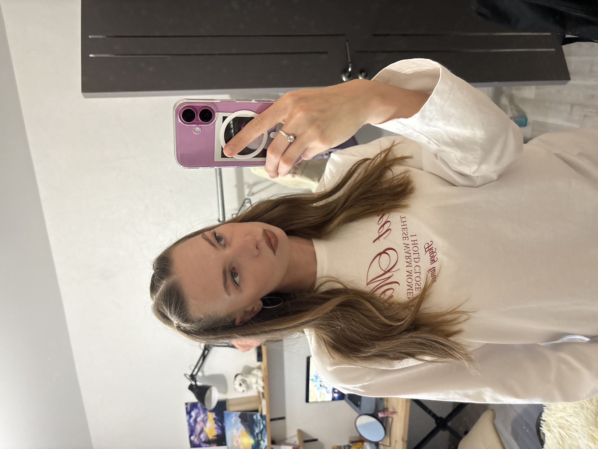 A young woman is taking a selfie in a room, holding a phone with a purple case. She is wearing a light-colored long-sleeved shirt with red text. The text on her shirt appears to be reversed, reading "MOMENTS CLOSE THESE" and "MOMENT THESE". She has long brown hair and is wearing a ring on her left hand. The room appears to be a bedroom or living space, with a desk and some artwork visible in the background. The lighting suggests it might be daytime, but there are no strong indicators of the time of day or weather. The overall mood is casual and personal.