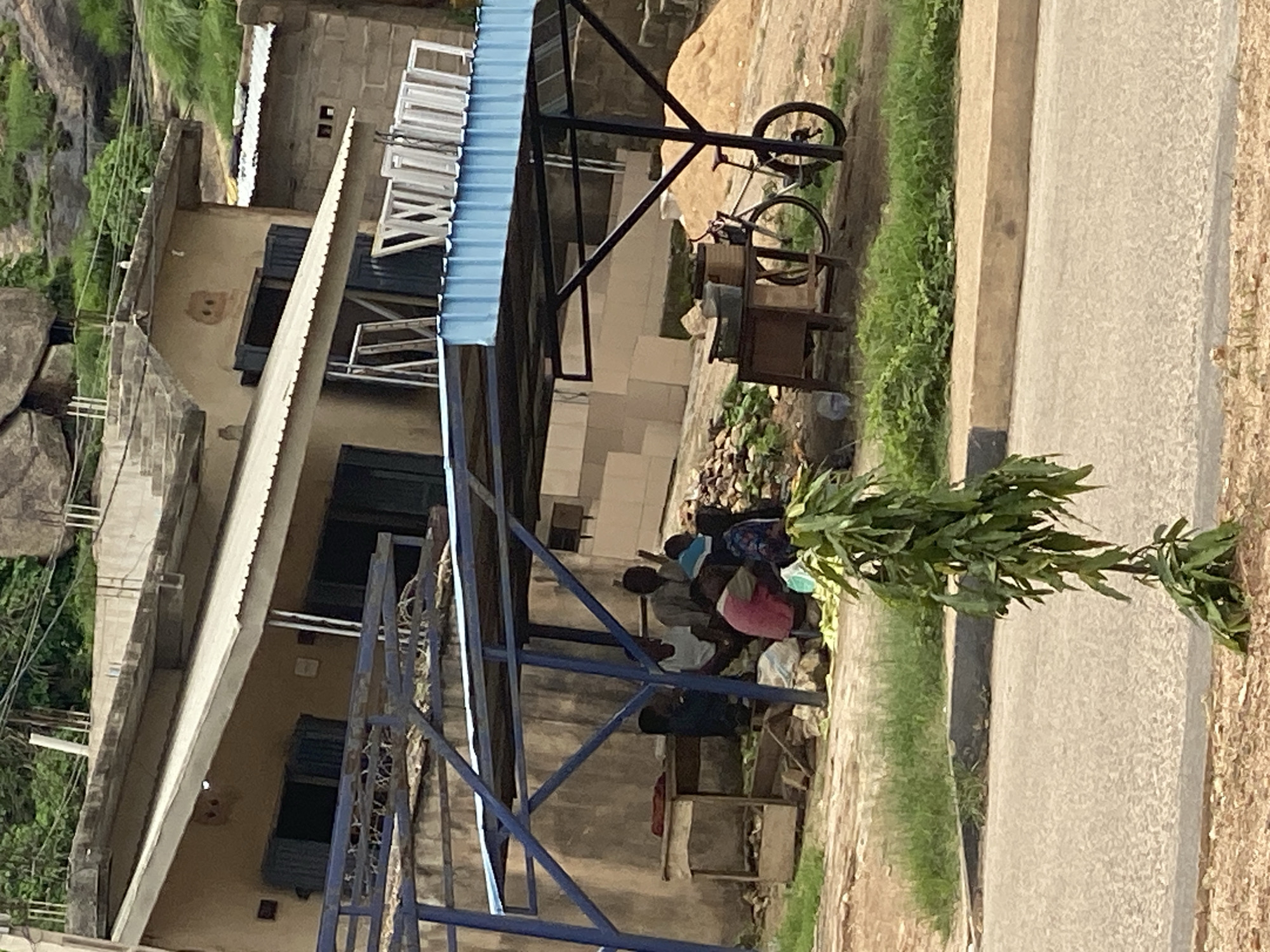 The image shows a group of people gathered under a blue metal structure with a corrugated metal roof, likely a shelter or market stall, in Gura Top, Nigeria. The scene appears to be outdoors during the daytime, with sunlight casting shadows.
Several individuals are visible, sitting or crouching. One person wears a pink garment, another a white shirt, and others are in darker clothing. They are positioned near a collection of what appear to be leafy green vegetables, suggesting a market setting or food preparation. A rustic wooden stall is present nearby.
In the background, a building constructed with concrete blocks and a corrugated metal roof is visible. Several windows with white frames are stacked outside the building. A bicycle is leaning against the structure, with its wheels visible. The overall environment suggests a rural or semi-urban area, with vegetation and rocky outcrops in the distance, consistent with the Gura Top location. The weather appears to be clear and sunny. There is no visible text in the image.