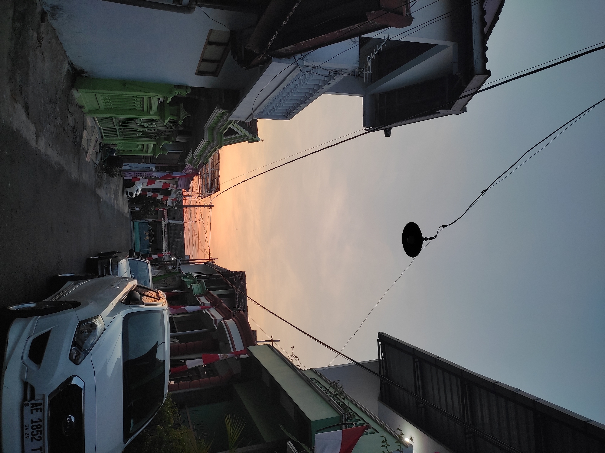 The image depicts a residential street in Ponorogo, Indonesia, during dusk. The sky is a gradient of pale blue and orange, indicating the setting sun. On either side of the narrow asphalt road are two-story houses with balconies and tiled roofs. Indonesian flags are displayed prominently on some of the houses, suggesting a celebration or national event.
Several vehicles are parked along the street. In the foreground, a white car is parked on the right side of the road, facing away from the camera. The license plate is partially visible and reads "AE 1852 T". Further down the street, another car, possibly silver, is parked behind the white car.
Overhead, electrical wires crisscross the sky, and a black lamp fixture is suspended from one of these wires, casting a shadow. The houses exhibit a mix of architectural styles, with some featuring ornate railings and green decorative elements. The ground on the left side of the street is a mixture of concrete and dirt. The overall atmosphere is tranquil, with the twilight casting long shadows and softening the edges of the buildings. There are no people visible in the frame.