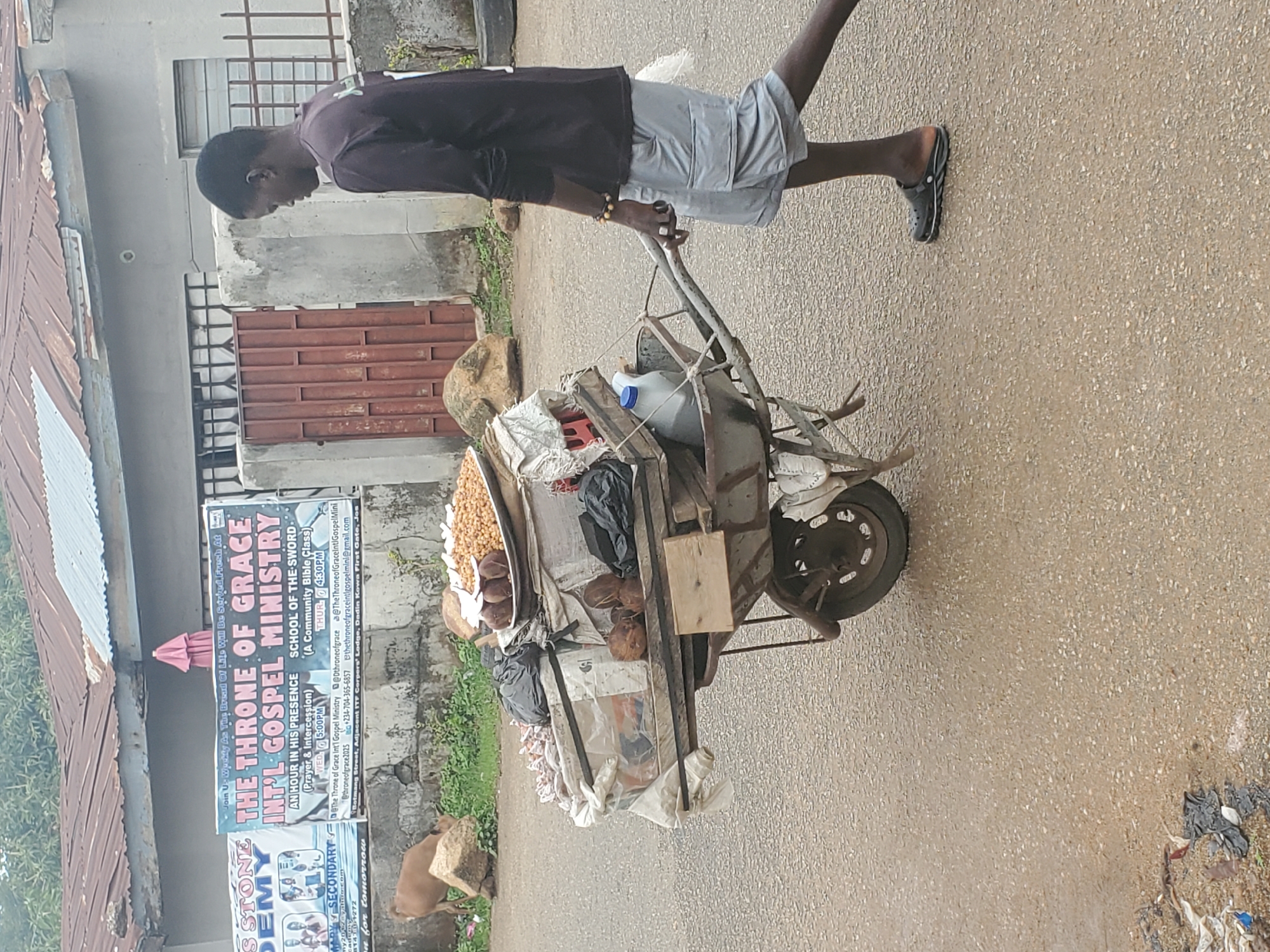 A man is walking along a street in Jos, Nigeria, pulling a heavily loaded handcart. The cart contains sacks, bags, coconuts, and other items. The man is wearing a dark long-sleeved shirt, light-colored shorts, and sandals. In the background, there's a building with a sign for "THE THRONE OF GRACE INT'L GOSPEL MINISTRY" and "STONE DEMY SECONDARY." The street appears to be paved. There are some dogs standing on the side of the street. The sign indicates the location is on Botn Street, Adjacent ITF Corpers' Lodge, Dadin Kowa First Gate, Jos.