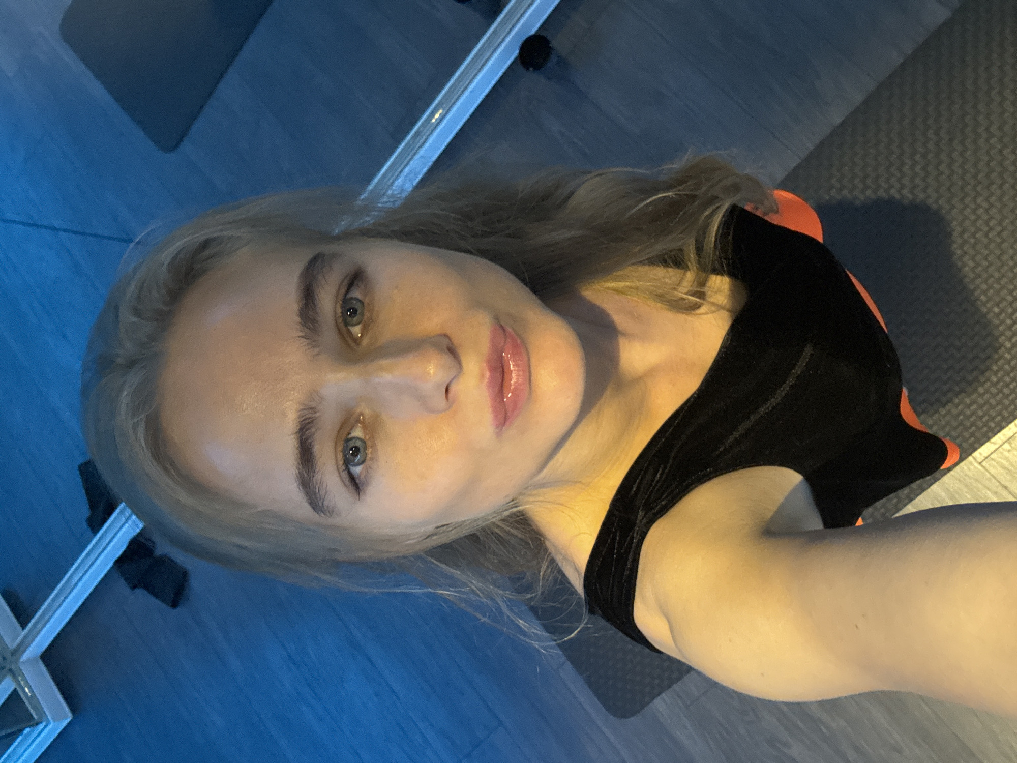 A woman with blonde hair, fair skin, and blue eyes is looking up at the camera. She is wearing a black leotard with an orange trim. She appears to be in a fitness studio, indicated by the presence of a grey exercise mat. The floor is blue. There's a reflective surface, potentially a mirror, visible along the edge of the frame. The location and city are unknown.