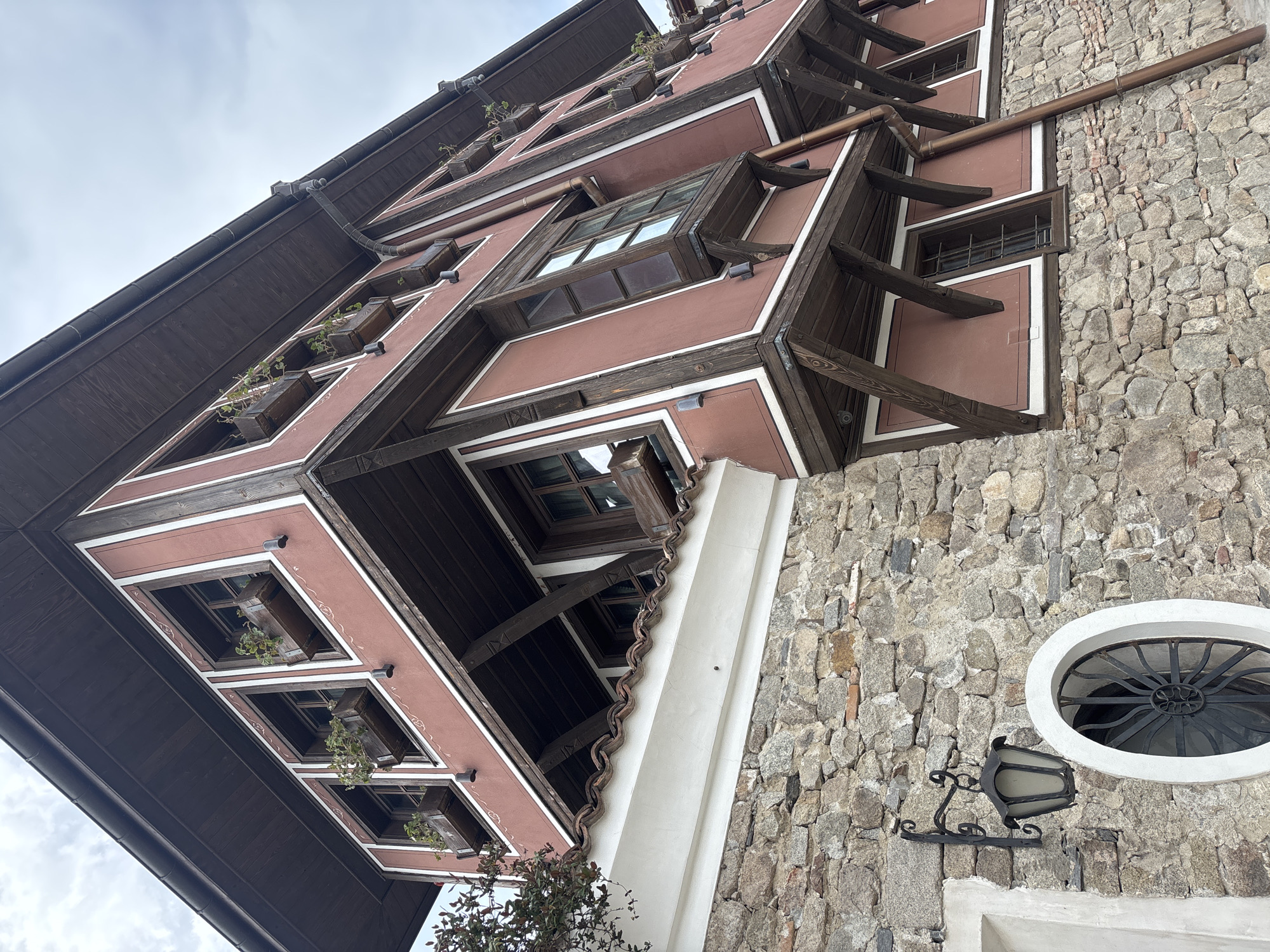 The image depicts the exterior of a historic building in Plovdiv, Bulgaria. The building has a distinctive architectural style with two visible sections. The left section is characterized by a maroon facade with white trim and dark brown wooden beams and window frames. Windows are recessed into the facade and some feature small planter boxes with greenery. The right section of the building is constructed of rough-hewn stone. A decorative black wrought-iron lamp is mounted on the stone wall, near a small oval window. The sky is visible in the upper left corner and appears overcast.