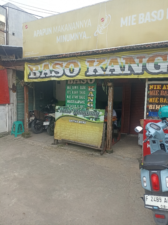 The scene is in front of a restaurant in Jatinangor, Indonesia, located at coordinates -6.933 latitude and 107.773 longitude. The restaurant is named "Baso Kangkung" and serves a variety of Indonesian noodle soup dishes, including "mie yamin baso", "mie kuah baso", and "mie ayam baso". The restaurant also sells "batagor kuah baso" and "batagor bumbu". A sign above the restaurant reads "Apapun Makanannya Minumnya..". A person is sitting at a table inside the restaurant. A teal-colored stool sits on a patch of dirt and grass. There is a motorcycle parked in front of the restaurant, facing towards the camera. The motorcycle is blue and has a license plate that reads "F 2489 AI". The street in front of the restaurant is paved and appears to be in a residential area.