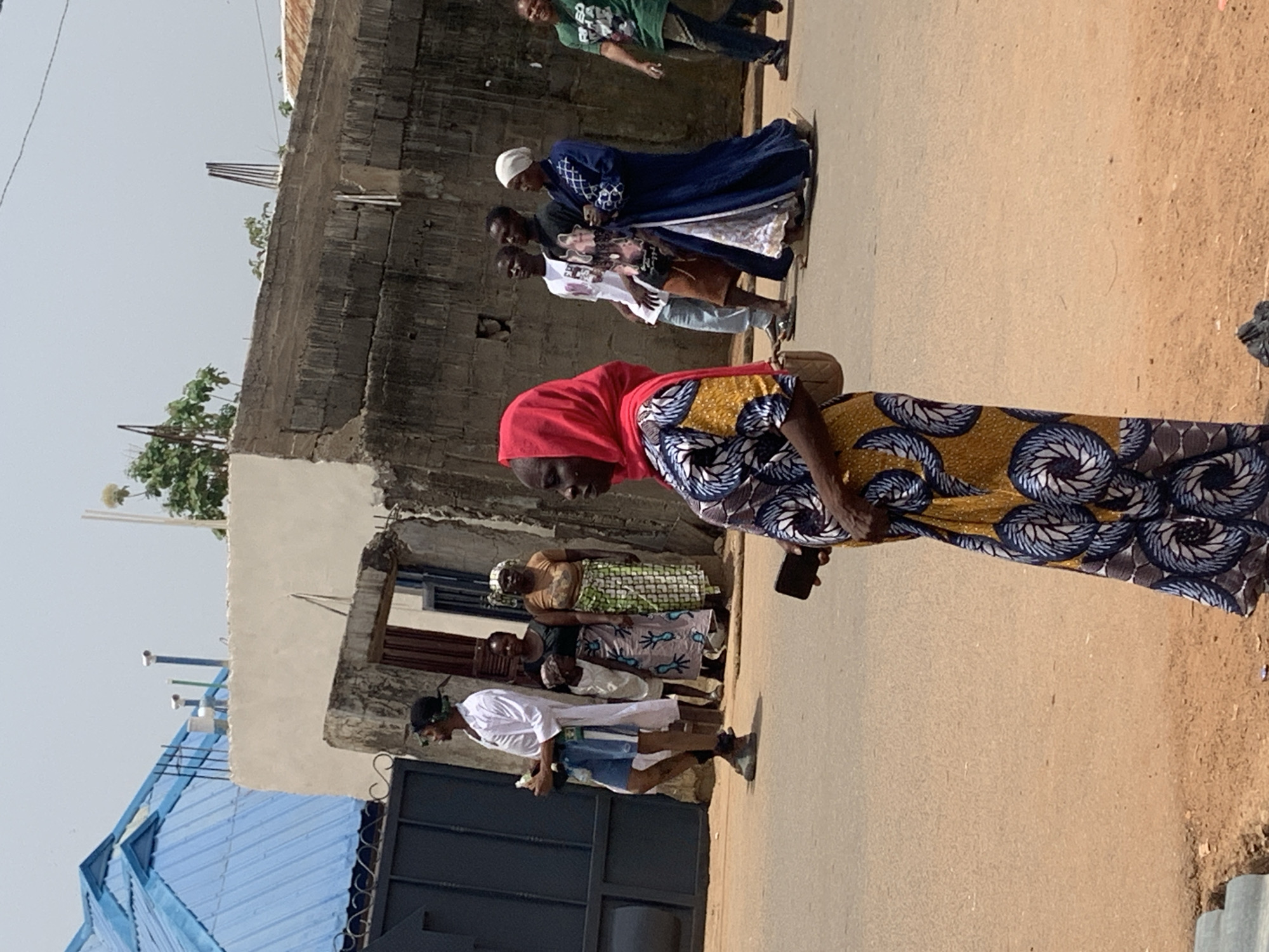 In Akwanga, Nigeria, a group of individuals are walking along a paved road adjacent to buildings. A woman wearing a red head covering and a patterned dress is prominent in the foreground, holding a dark-colored object. Behind her, other people are walking in the same direction, some wearing traditional clothing. The buildings on the side of the road vary in construction, with one having a blue roof and another constructed of stone. The environment is sunlit, casting shadows. The road surface is light brown.