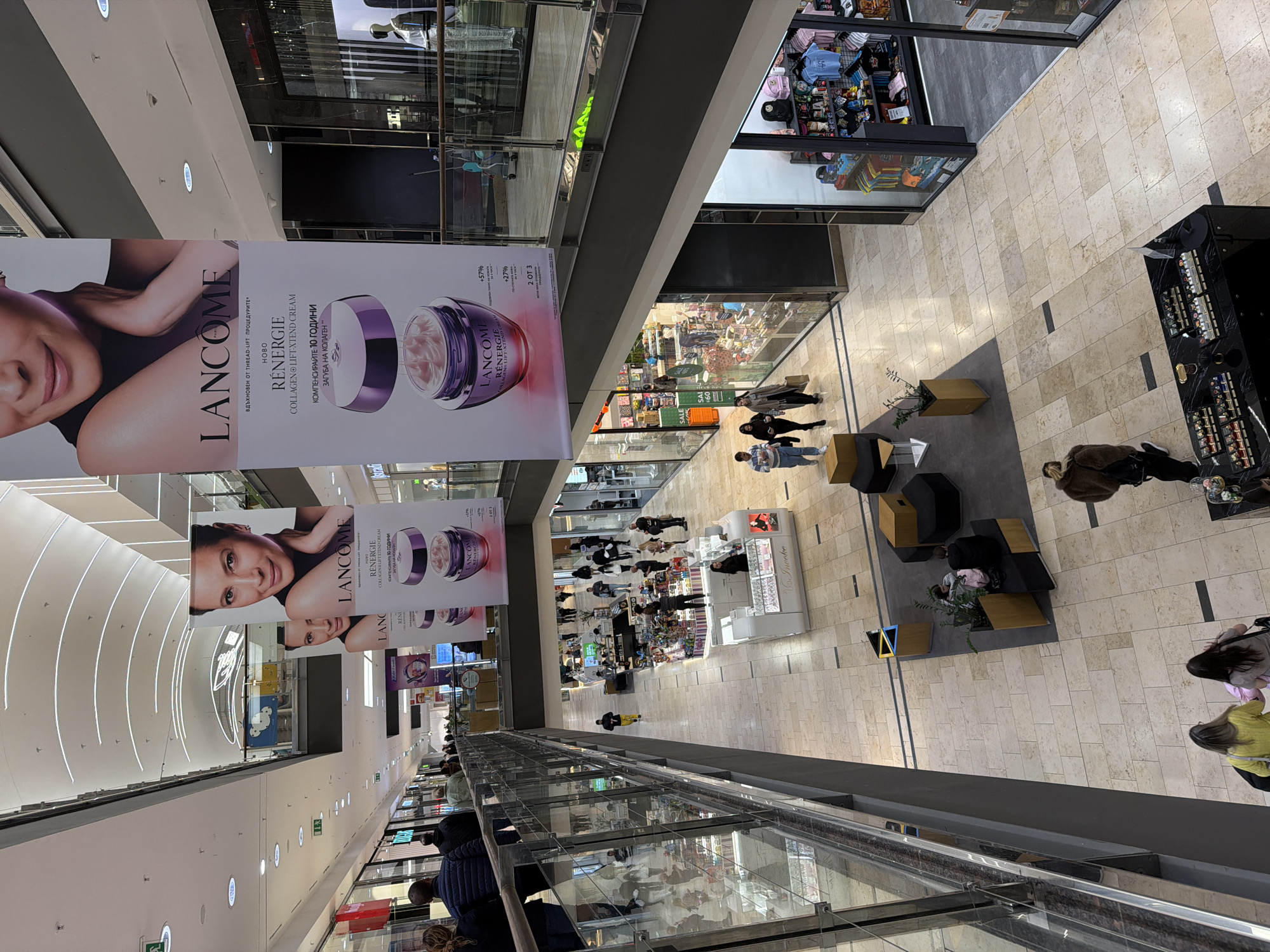 Captured from an elevated perspective, the image shows an interior view of a shopping mall, possibly in Sofia, Bulgaria, given the presence of Bulgarian text on the Lancôme advertisements. Banners displaying Lancôme advertisements featuring close-up images of women and product promotions in Bulgarian hang from the ceiling of the mall. Shoppers meander through the corridors, some pausing near retail displays or seated in the lounge areas featuring black and gold seating. The architecture of the mall includes multiple levels with glass railings. Storefronts line the walkways. The floor is covered in light-colored stone tiles. The mall appears well-lit with rows of ceiling lights visible in the upper left.