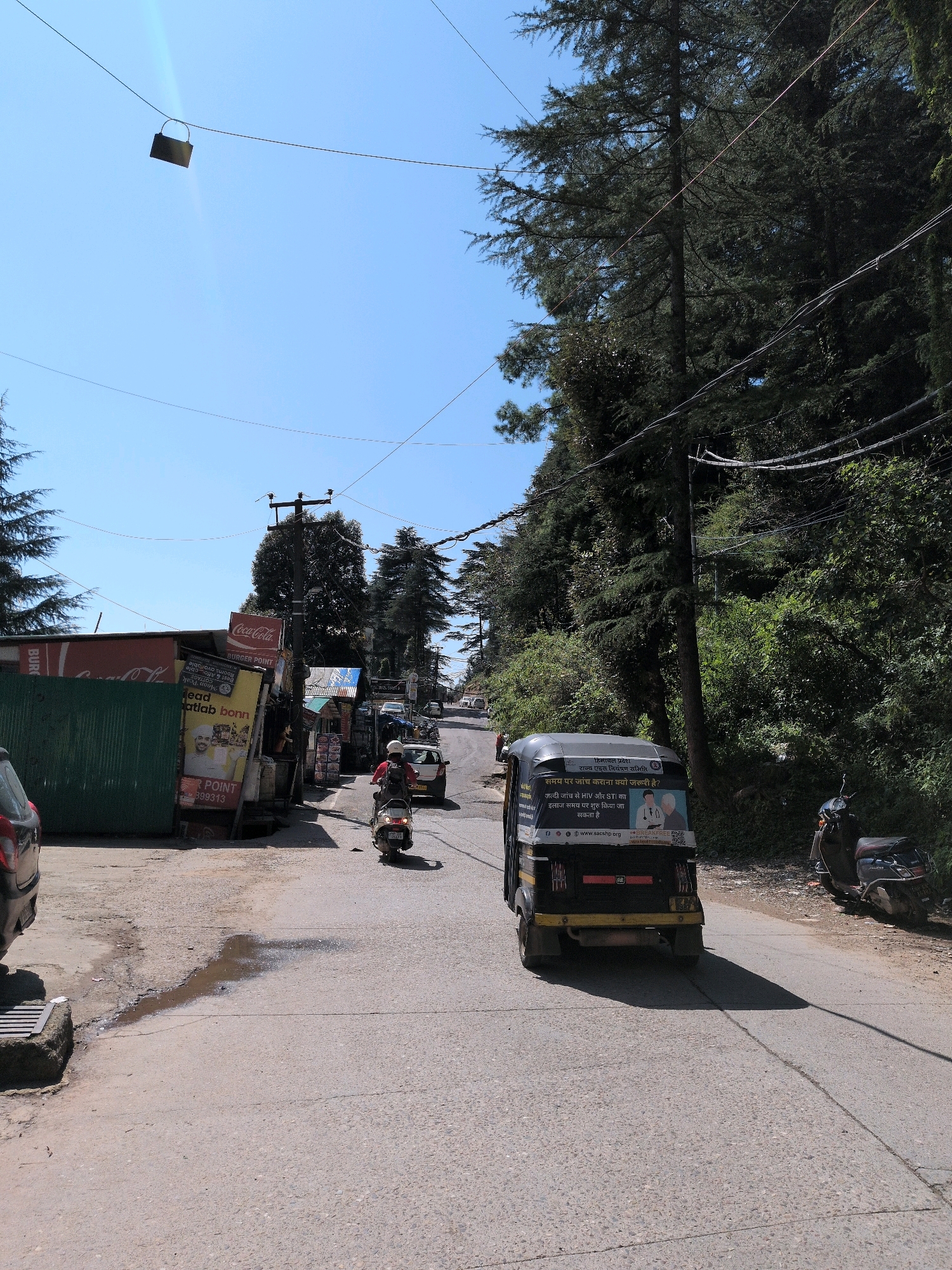 A wide-angle, eye-level, vertical shot captures a street scene in Bhagsu, India, on a bright, sunny day. A paved road with some water puddles curves uphill, flanked by dense, dark green pine trees and various buildings on the left. The sky is clear blue with a slight lens flare visible in the upper left quadrant. Overhead, power lines crisscross the frame, and a dark rectangular object, possibly a small lock, hangs from one of them.
In the foreground on the left, the rear of a dark car is partially visible. Adjacent to it is a green corrugated metal wall, behind which are several advertisements, including a large Coca-Cola sign. A few small shops with awnings are visible further up the street on the left.
The middle ground features a busy street with traffic. A person on a light-colored scooter, wearing a white helmet and a red shirt, is moving uphill. Further up, a white car is visible. Dominating the center of the road is a three-wheeled auto-rickshaw, painted black and yellow, displaying a poster with text in Hindi and two images of elderly individuals. The auto-rickshaw has a visible license plate and is casting a long shadow on the road.
On the right side of the road, more dense pine trees form a natural backdrop. A dark-colored scooter is parked on the shoulder of the road, with a person on it. The overall impression is a bustling, somewhat rustic mountain town street during daytime.